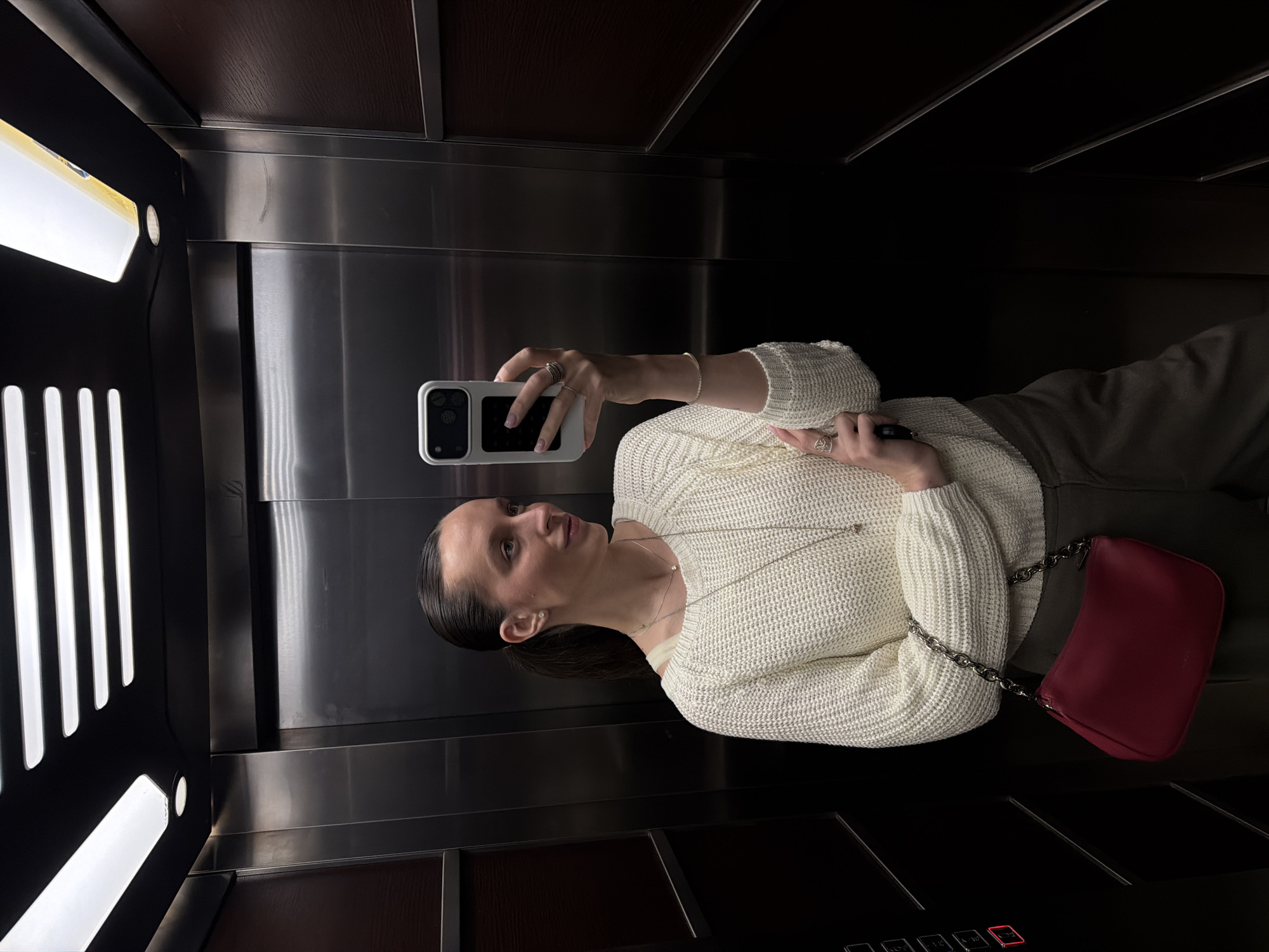A woman is taking a selfie inside an elevator. She is fair-skinned, wearing a white knit sweater, grey pants, a silver necklace, and several rings. She is holding a white smartphone with a black screen. A red purse with a silver chain hangs from her shoulder. The elevator has stainless steel walls and a dark brown ceiling, along with a panel of buttons with numbers and a red button. A light fixture is on the left wall. The location is not discernible from the given details.