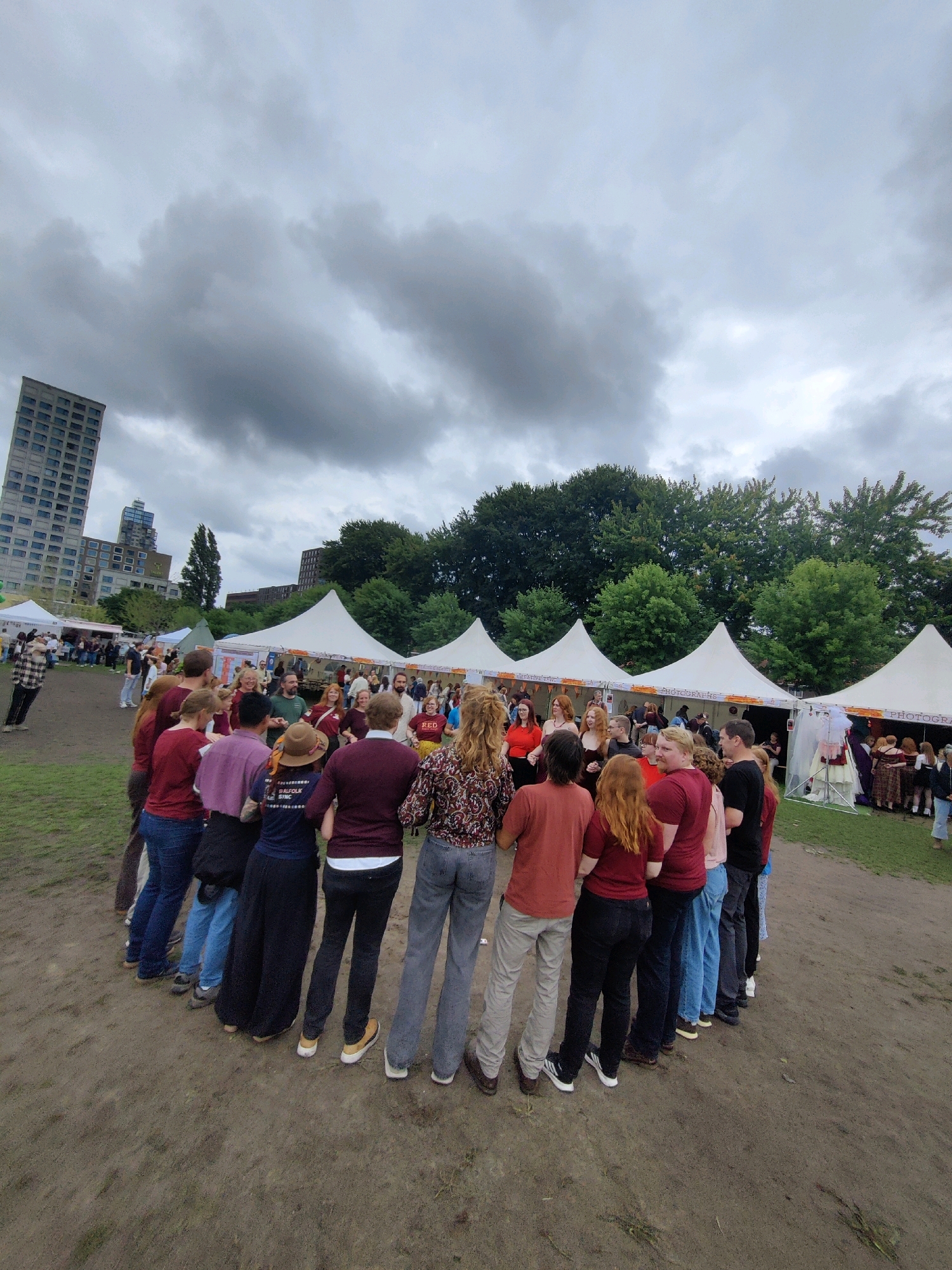 A group of approximately 30 people are standing in a circle on a dirt ground, facing inward. The individuals are dressed in casual clothing, with some wearing t-shirts and jeans, others wearing skirts and dresses. The event appears to be outdoors, with several white tents set up in the background. Beyond the tents, trees and buildings are visible, including a tall, modern high-rise building with many windows. The sky is overcast with gray clouds. The scene takes place in the city of Tilburg, Netherlands, potentially at a festival or market given the tents and the gathering of people.