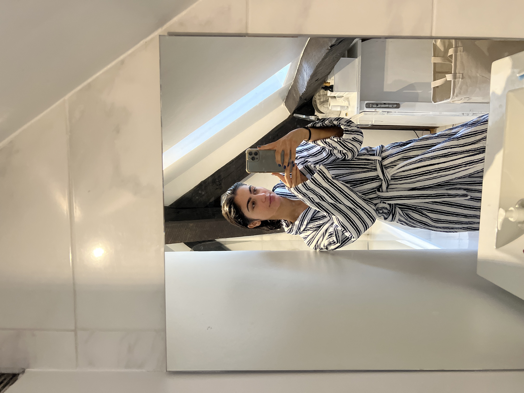 A young woman is taking a mirror selfie in a bathroom. She is wearing a black and white striped robe and has wet hair. She is holding a smartphone in her right hand and is looking at her reflection. The bathroom appears to be in a Parisian apartment, as evidenced by the sloped ceiling with exposed wooden beams and the style of the tiled walls, common in older Haussmannian buildings. On a shelf in the background, there are several small bottles and containers, possibly skincare or beauty products, and a beige laundry basket. The lighting suggests it is daytime.