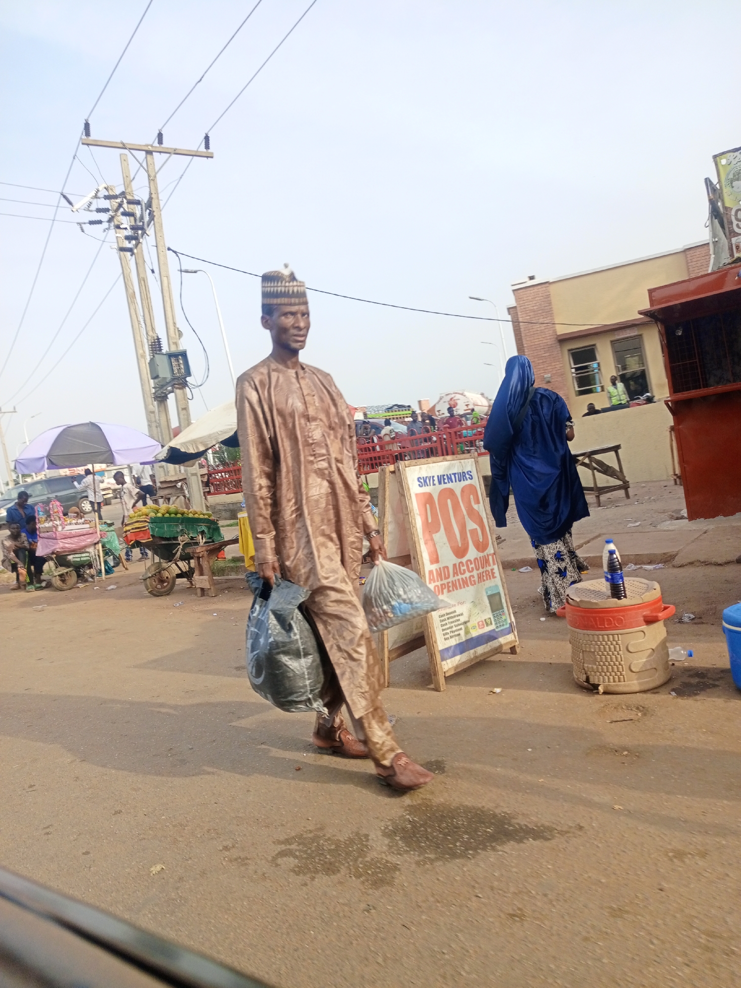 The image, taken in Keffi, Nigeria, depicts a man dressed in traditional Northern Nigerian attire, consisting of a brown, flowing tunic and trousers, and a patterned cap, walking on a paved street. He carries two dark-colored bags in his hands. A woman wearing a blue hijab stands near a sandwich board advertising "SKYE VENTURES POS" with details for cash deposit, cash transfer, and account opening. A cooler labeled "RONALDO" with water bottles sits nearby. In the background, there are market stalls with vendors and customers, and a building with brick accents. Overhead power lines run across the sky. The scene captures a typical street scene in the city, blending commerce and daily life.