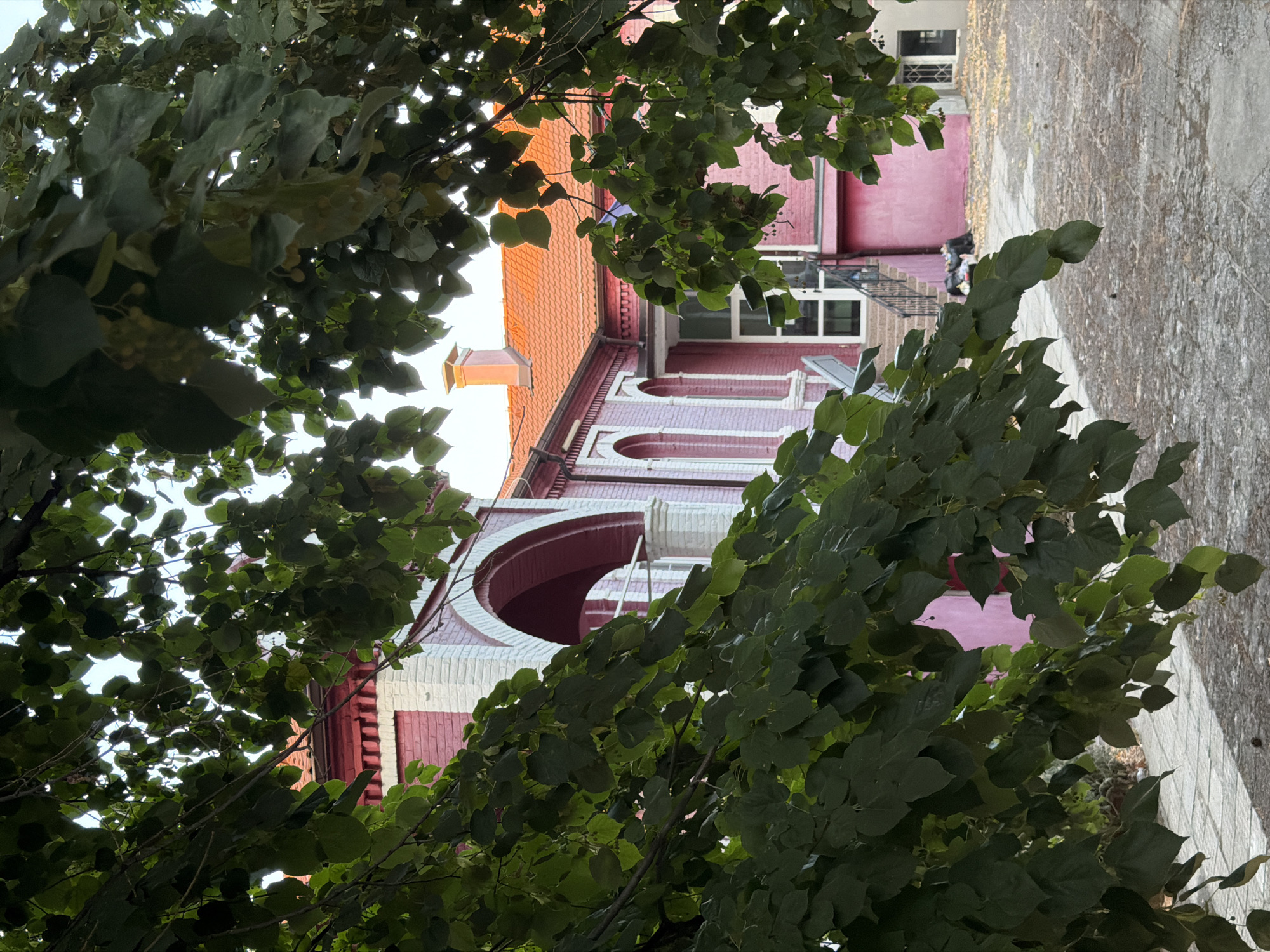 An outdoor shot shows a pink and red brick building with a terracotta tiled roof in Svilengrad, Bulgaria. The building has arched doorways and windows. Lush green foliage from trees hangs in the foreground, partially obscuring the view of the building. The ground in front of the building is paved with stone and there are several black trash bags piled up near a set of stairs. The sky is a pale blue and appears to be slightly overcast.