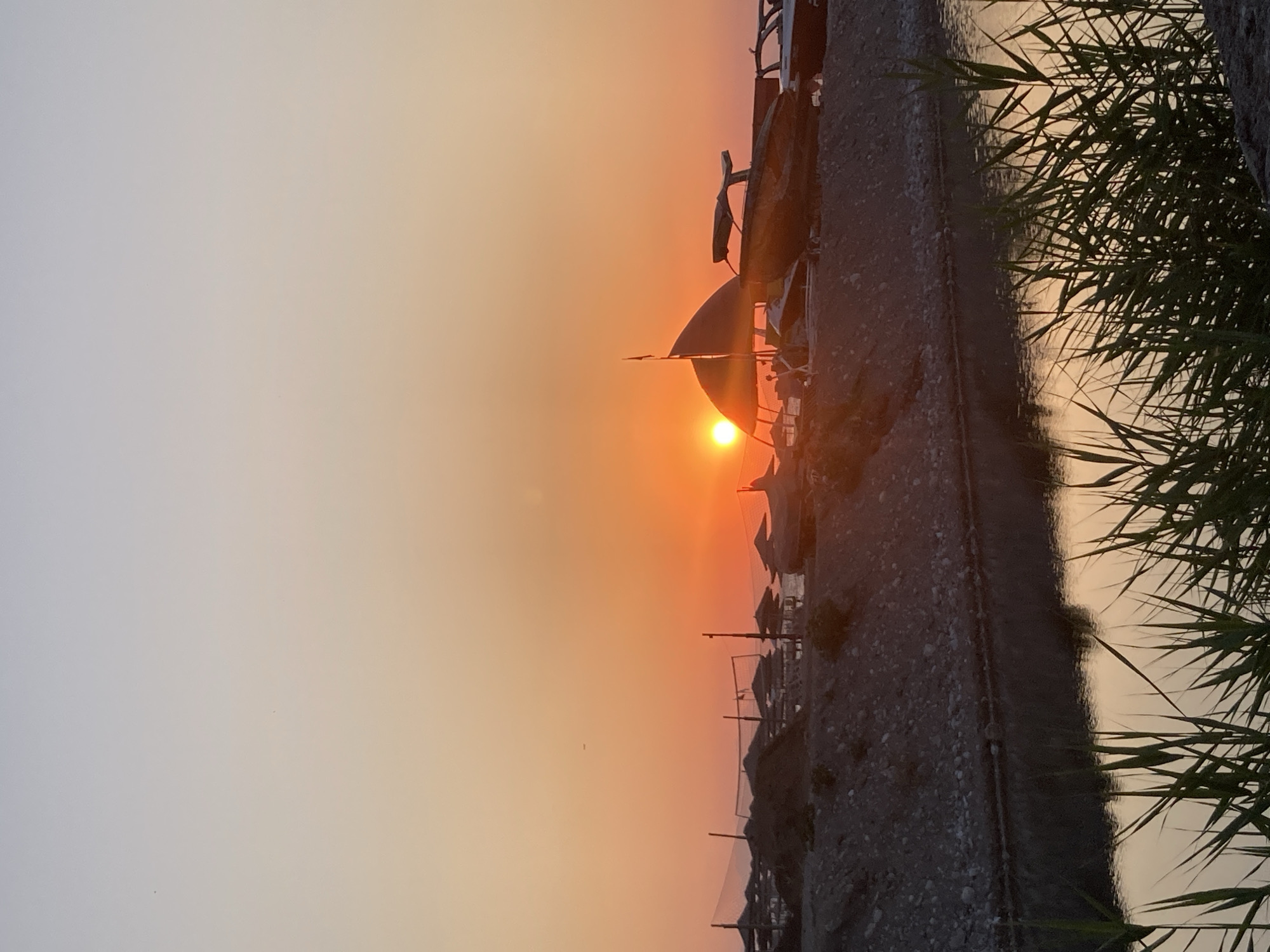 The image captures a serene sunset scene in Kemer, Turkey, likely near a coastal area. The sky transitions from a pale blue at the top to vibrant orange and yellow hues near the horizon, where the sun is setting. In the foreground, tall green reeds and grasses are silhouetted against the water. A calm body of water reflects the warm colors of the sunset.
Beyond the water, a rocky embankment rises, topped with a collection of beach umbrellas, some appearing to be under construction or not in use. Beach volleyball nets are visible among the umbrellas. Several boats, including speedboats and a larger vessel with a sail-like structure, are parked on the shore or in the shallow water. The overall atmosphere is peaceful, with the natural beauty of the sunset enhanced by the elements of a coastal resort. There are no discernible people or actions taking place in the image.