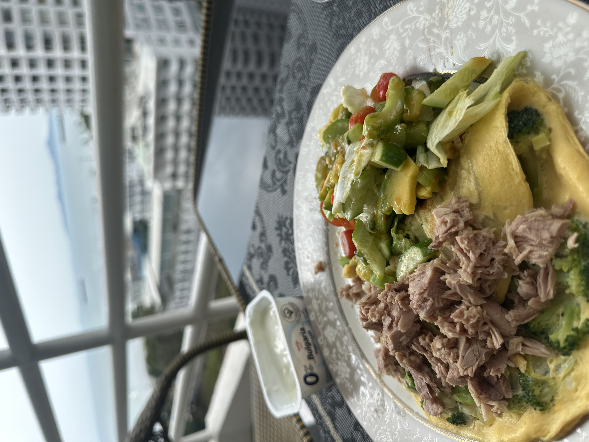 The image shows a plate of food on a table in Pattaya City, Thailand. The food consists of a salad with avocado, cucumber, and tomatoes, alongside an omelet with broccoli and tuna. A small container of Bulgaria yogurt is also visible on the table. The background shows a blurred view of high-rise buildings and the ocean through a balcony railing. The table has a patterned tablecloth, and the plate is white with a floral design. The setting suggests a meal being enjoyed on a balcony with a view of the city and coastline.
