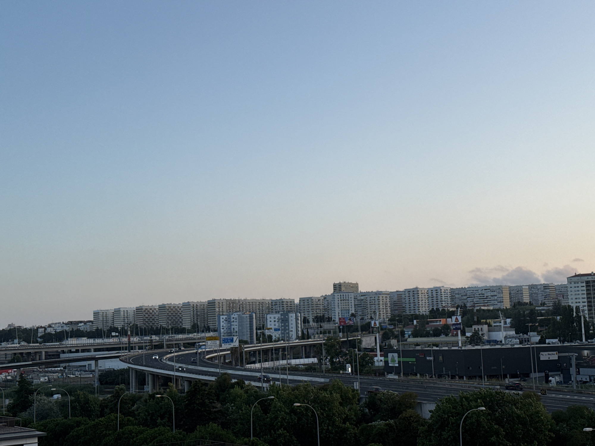 The image shows a panoramic view of the city of Loures, Portugal, likely taken from an elevated position. The scene is dominated by a complex highway interchange with multiple levels of ramps and bridges, curving through the landscape. Several cars are visible on the highway, indicating moderate traffic. In the background, a dense collection of residential buildings, characterized by their uniform, modern architecture, stretches across the horizon. These buildings appear to be multi-story apartment blocks. Vegetation, including numerous trees and some bushes, is present in the foreground and around the highway, suggesting a green urban environment. The sky is clear with a soft, gradient blue, possibly indicating early morning or late afternoon light. Some clouds are faintly visible near the horizon. The overall impression is one of a bustling urban area with significant infrastructure, set against a backdrop of residential development.