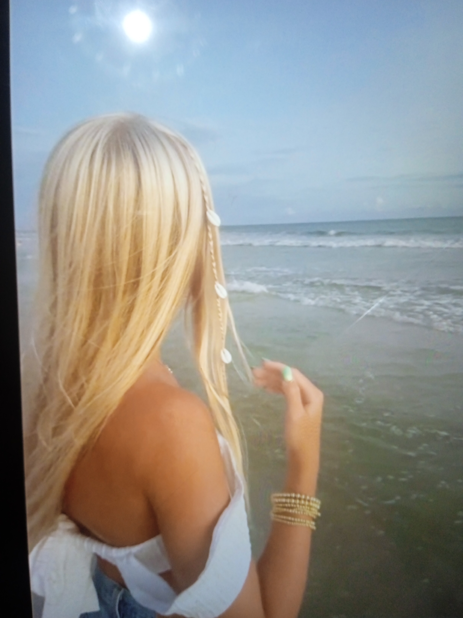 The image shows a woman with long blonde hair, viewed from behind, standing on a beach. Her hair is partially braided and adorned with small white shells. She is wearing a white off-the-shoulder top and denim shorts. Her right arm is extended, and she is wearing a gold bracelet on her wrist. The ocean is visible in the background with waves breaking on the shore under a clear sky. The sun is high in the sky, creating a bright reflection. The image quality is low, with a visible sheen and pixelation. There are no discernible landmarks or elements within the image that would indicate the city of Chernivtsi, Ukraine, or any other specific urban location. The scene is entirely natural and coastal.