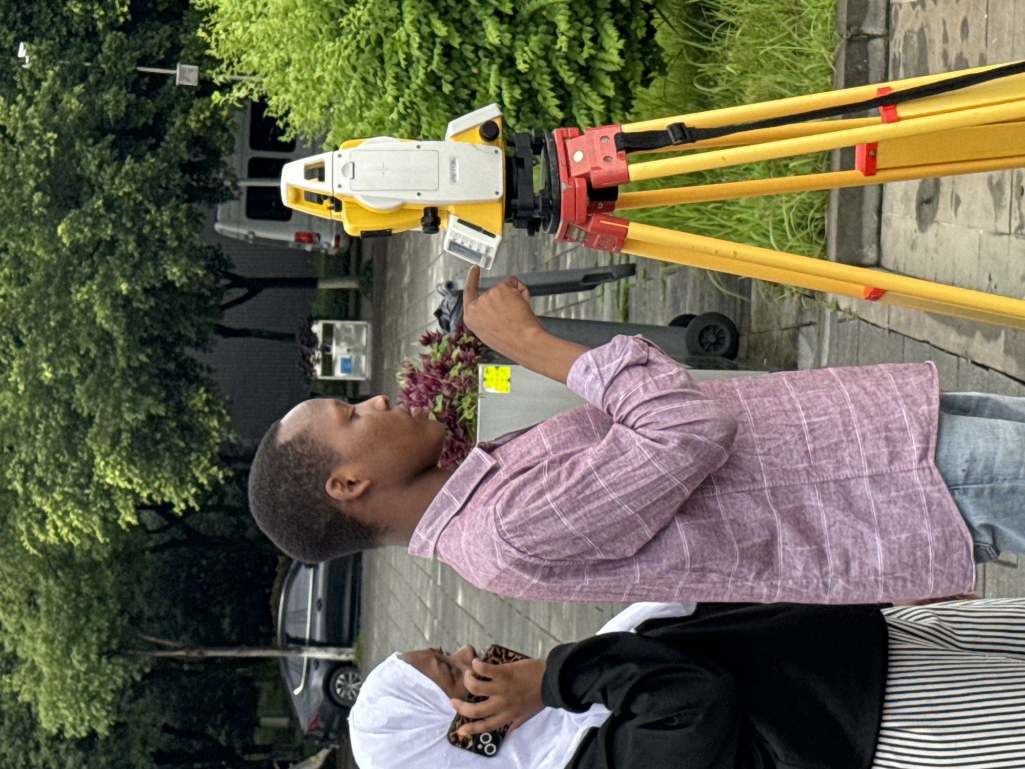 Two young individuals, a female wearing a white hijab and a black jacket over vertically striped pants, and a male wearing a pink checkered shirt and blue jeans, are outdoors. The female is holding a smartphone to her ear, while the male is pointing his right index finger towards a surveying instrument. The surveying instrument, a yellow and white total station, is mounted on a red and yellow tripod. The scene appears to be on a paved area with some greenery and trees in the background, suggesting an urban or suburban park setting. A silver car and a white van are visible in the background, as well as a trash receptacle. The overall atmosphere suggests an educational or professional activity related to surveying. This image was captured in Jiangjin, China.