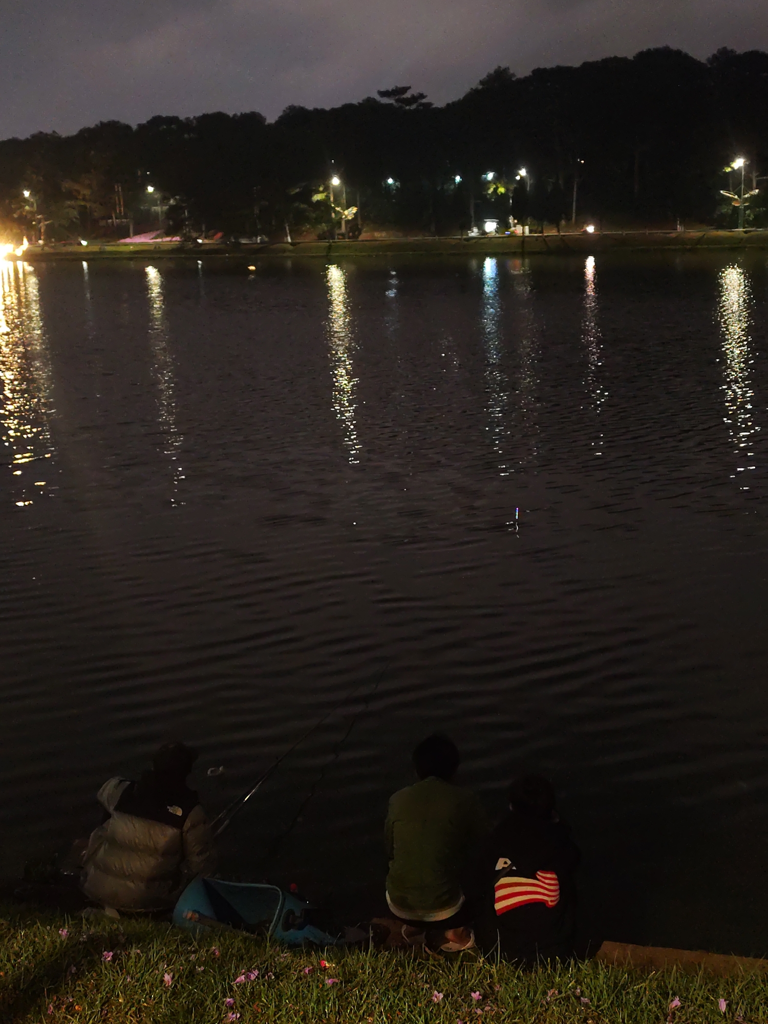 At night, on the shore of Xuan Huong Lake in Da Lat, Vietnam, three individuals are fishing. Two are seated close together, while another is slightly separated. A fishing rod is visible near the person on the left. The lake reflects lights from the distant shore, and a lighted fishing float is visible in the water. Grass and some flowers are at the edge of the lake.