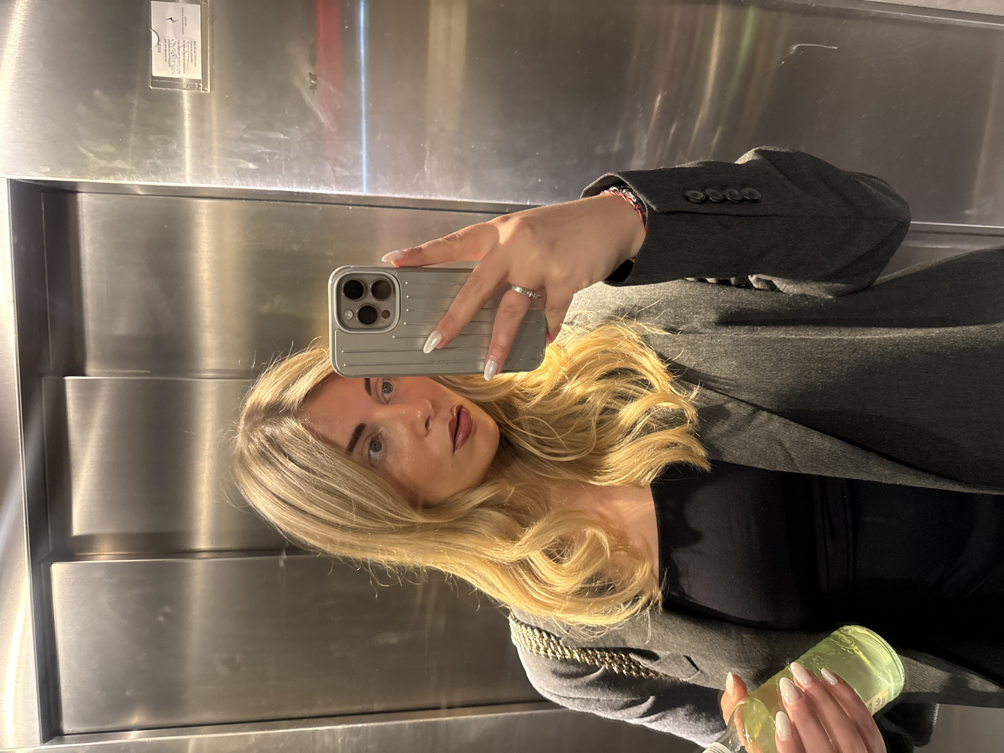 A young woman is taking a selfie in what appears to be an elevator. She is holding a silver-colored smartphone in her right hand, and in her left hand, she holds a lime-green glass bottle. The woman has shoulder-length blonde hair and wears a dark, formal jacket over a black top. She also has a ring on her finger. The elevator walls are metallic and have a reflective surface. A small sign is affixed to the upper section of the wall. The scene is enclosed and relatively confined, indicating the interior space of the elevator. While a specific location within Paris is not discernable, the presence of an elevator suggests an indoor setting in an urban building in Paris, France.