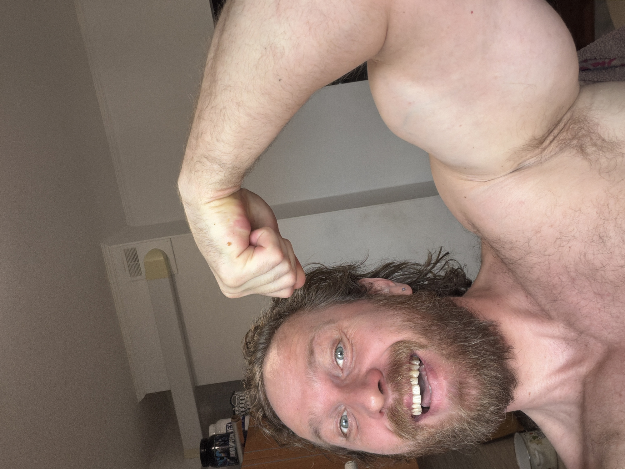 A close-up, angled shot features a smiling man with a thick beard and long hair, flexing his right bicep. He has light skin and blue eyes. His teeth are visible, and there is some visible wear on them. The background is a simple interior with a vent on the white wall. A cabinet with some jars on top is partially visible. The setting is Saint Petersburg, Russia.