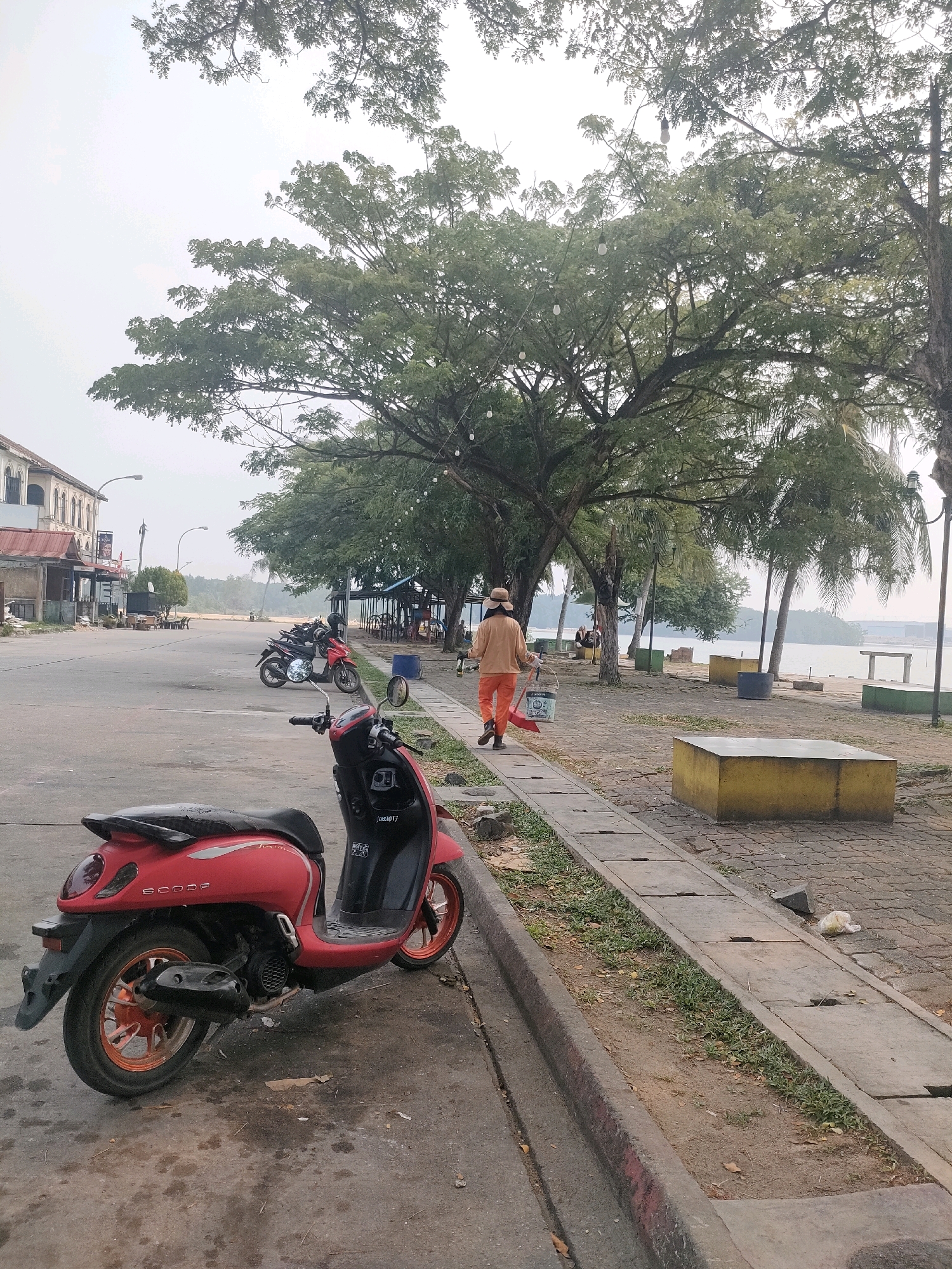 A woman in an orange outfit and a hat walks away from the camera on a concrete sidewalk next to a street. She is carrying a bucket in her left hand and a red object, possibly a shovel, in her right hand. The street is lined with several parked motorcycles, including a red scooter in the foreground. Across the street, there are buildings with awnings. Beyond the sidewalk and street, there is a body of water with trees and palm trees along the shore. The sky is overcast. This scene is located at Batam City, Indonesia, specifically along a waterfront area likely near a commercial district.