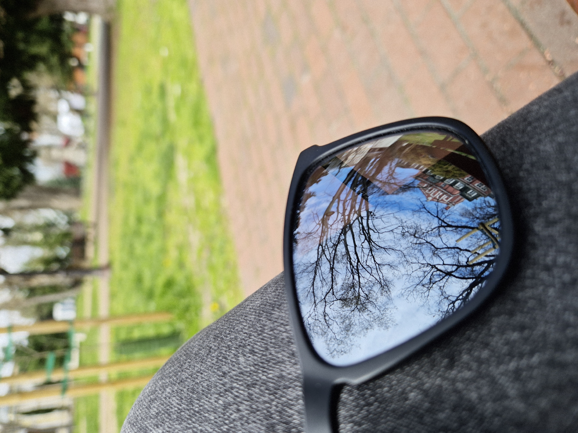 The image depicts a pair of sunglasses resting on a gray fabric surface, likely a person's leg. The lenses reflect a distorted view of a building with red brick walls and a blue sky with bare tree branches. The out-of-focus background suggests a park-like setting with green grass, a yellow wooden fence, and brick pavement. Given the architectural style and general environment, the location is likely in the city of Varna, Bulgaria, but this determination cannot be done forensically. No people are directly visible in the image, however their presence is implied by the sunglasses on a person's leg. The interaction is between the person and the environment, as captured by the reflection in the sunglasses.