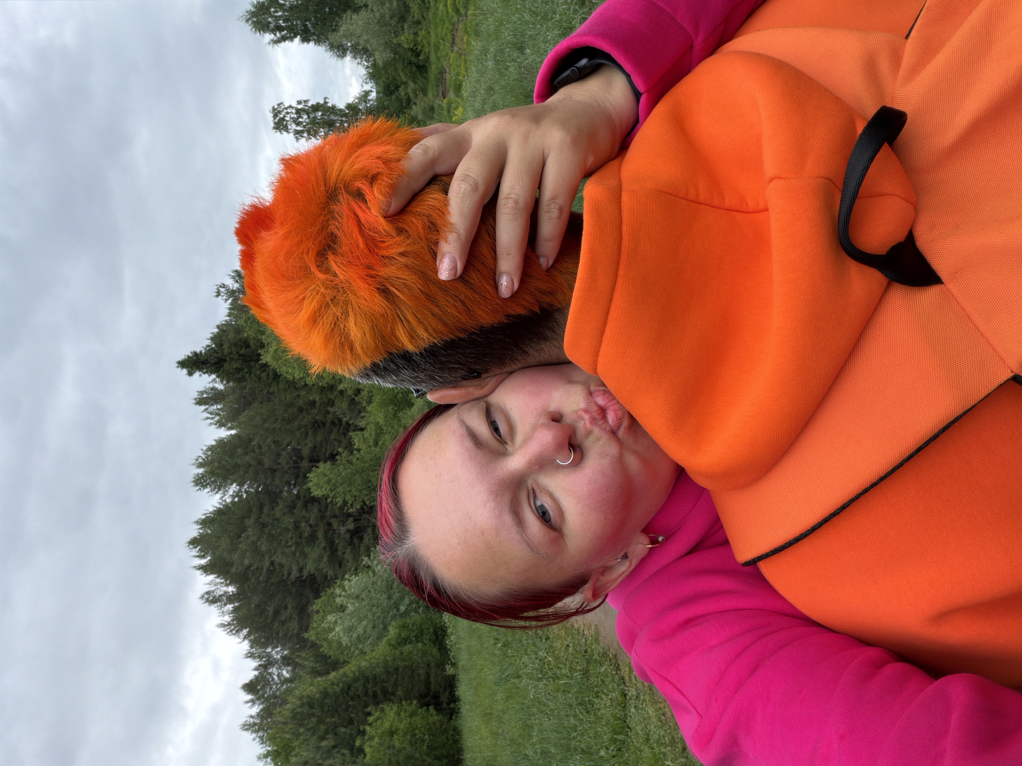 The photo is of a person with dyed pink hair and a nose ring, wearing a pink hoodie, holding an orange and black animal, possibly a dog, in their arms. The person is making a kissing face. The background is a green field and trees under a cloudy sky. The location may be in Chaikovsky, Russia.