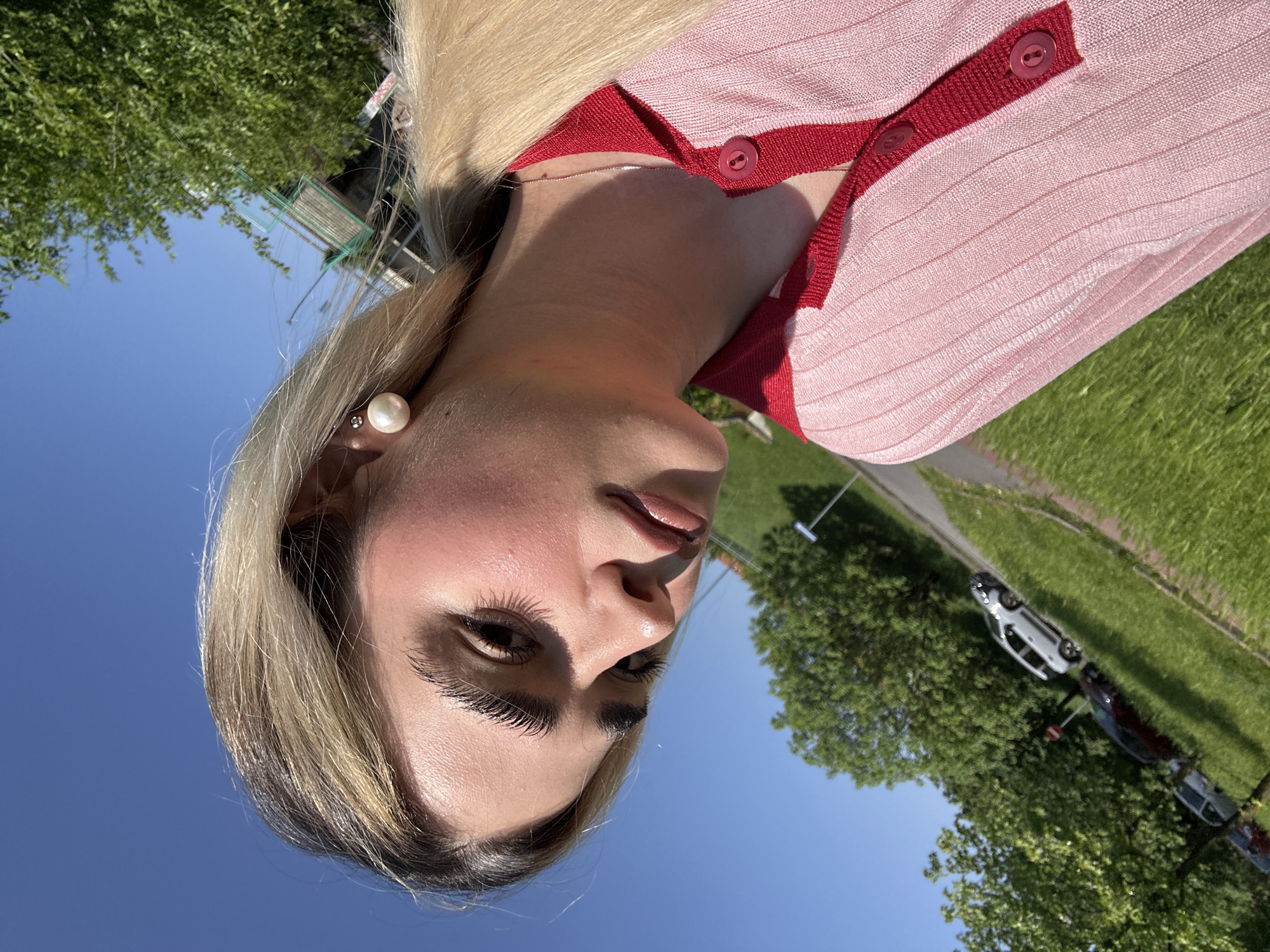 A woman with blonde hair and a pink and red cardigan poses for a photo outdoors. She wears a pearl earring in her left ear. The background consists of a blue sky, trees, grass, a sidewalk, and parked cars. The location is consistent with an outdoor setting in a city, possibly Milan based on the prompt.