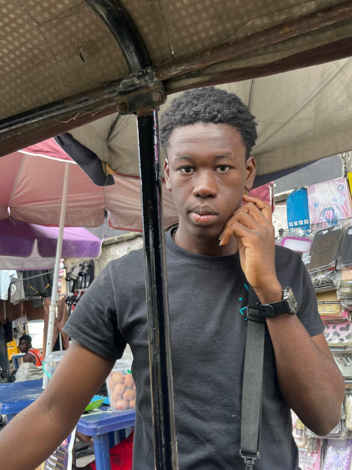 A young Black male with short, dark, curly hair stands in front of a market stall in Ado, Nigeria. He is wearing a black t-shirt and has a dark strap across his chest, possibly from a bag. His right hand is raised to his face, touching his cheek. He wears a watch on his left wrist. Behind him, the market stall is stocked with various items, including phone cases and what appear to be small, round fruits in a clear container on a blue table. Umbrellas, one pink and one purple, are visible in the background, as is clothing hanging on racks. Another person, a Black male wearing an orange vest, is seated in the background. The overall scene depicts a bustling street market environment.