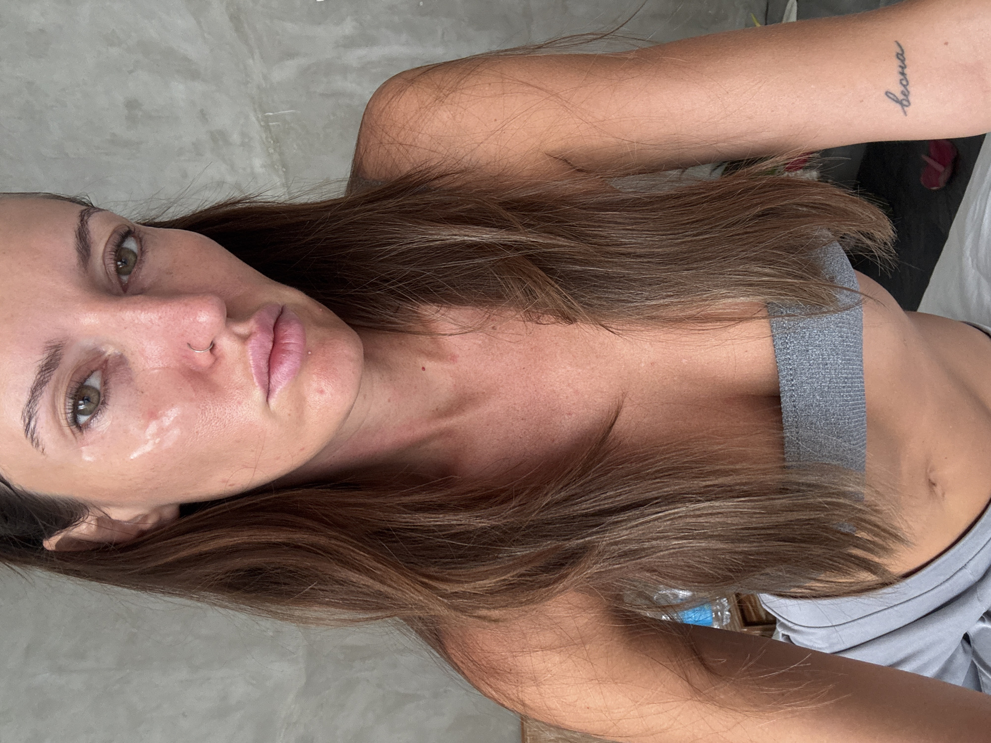 A woman with brown hair lies on a gray surface. She wears a gray top. There's a tattoo of a word on her arm. The environment appears to be a room with a gray wall. No confirmation on the exact location in North Kuta, Indonesia can be ascertained from this image.
