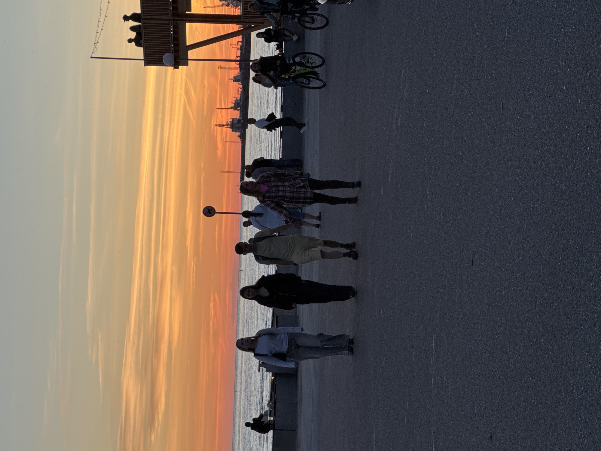 This is a photograph taken during sunset, likely on a promenade or waterfront in Tallinn, Estonia. The sky is filled with vibrant orange and yellow hues, indicative of a sunset. In the foreground, there is an asphalt surface. Several people are visible.
On the left side of the image, three individuals are walking towards the viewer. The leftmost person is a woman wearing a light-colored top and grey pants. Next to her is another woman in dark clothing. The third person is a man wearing a light-colored t-shirt and shorts, carrying a backpack.
Further to the right, a woman in a checkered shirt and black leggings is walking. Behind her, a person is riding a green bicycle. To the far right, another person is on a black bicycle. In the background, there are more people standing and sitting, and some are silhouetted against the bright sky.
The middle ground features the sea, with industrial structures and cranes visible on the horizon, suggesting a port area. On the right side of the frame, there is a raised wooden structure with people standing on it, possibly a viewing platform or a part of a building. A string of lights is visible above this structure. A pole with a circular sign depicting a dog is also present. The overall scene depicts a relaxed, evening atmosphere with people enjoying the sunset.
