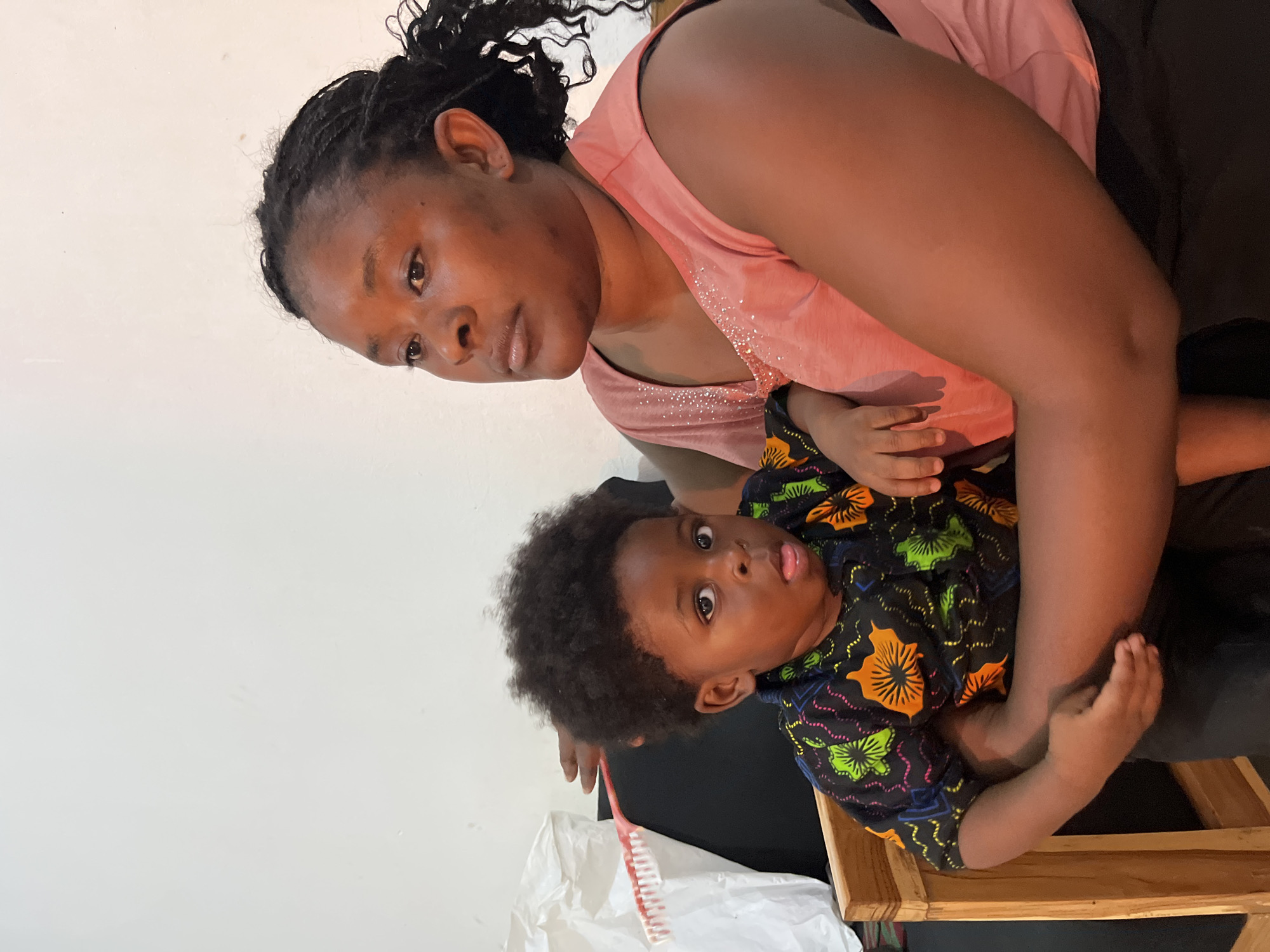 In Gura Top, Nigeria, a woman with dark skin and braided hair is holding a young child in her lap. The woman is wearing a pink sleeveless shirt with a shimmering neckline. The child has dark, curly hair and is wearing a colorful shirt with a floral pattern in green, orange, and blue. The child's mouth is slightly open, and their tongue is visible. They are sitting in a wooden chair. In the background, a white plastic bag and a black surface are visible. The wall behind them is plain and white. A red and white toothbrush is also visible in the background, positioned behind the child's head.
