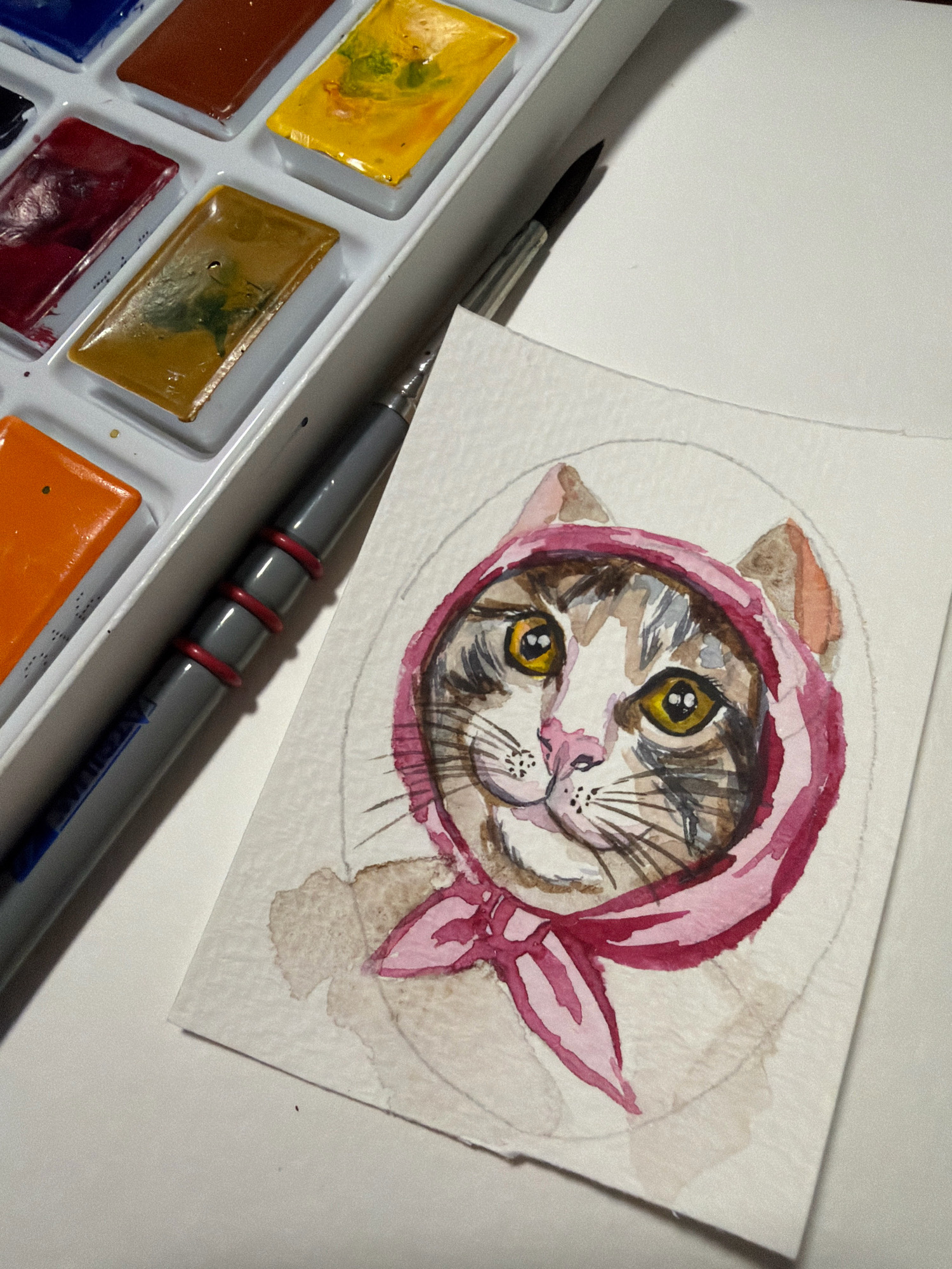 An overhead shot shows a watercolor painting of a cat wearing a pink headscarf and a watercolor paint palette. The painting is on a white paper and is on the right side of the frame, with a paint palette on the left. The cat's face is the focal point of the painting, with its yellow eyes and pink nose clearly visible. The cat has brown and black stripes on its fur, and its ears are peeking out from the headscarf. The paint palette is a white rectangular tray with several pans of watercolor paints. The paints are various colors, including blue, red, yellow, and orange. A paintbrush is also visible in the image, resting on the paint palette. The scene is lit from above, casting shadows on the paper and paint palette. The background is a plain white surface.