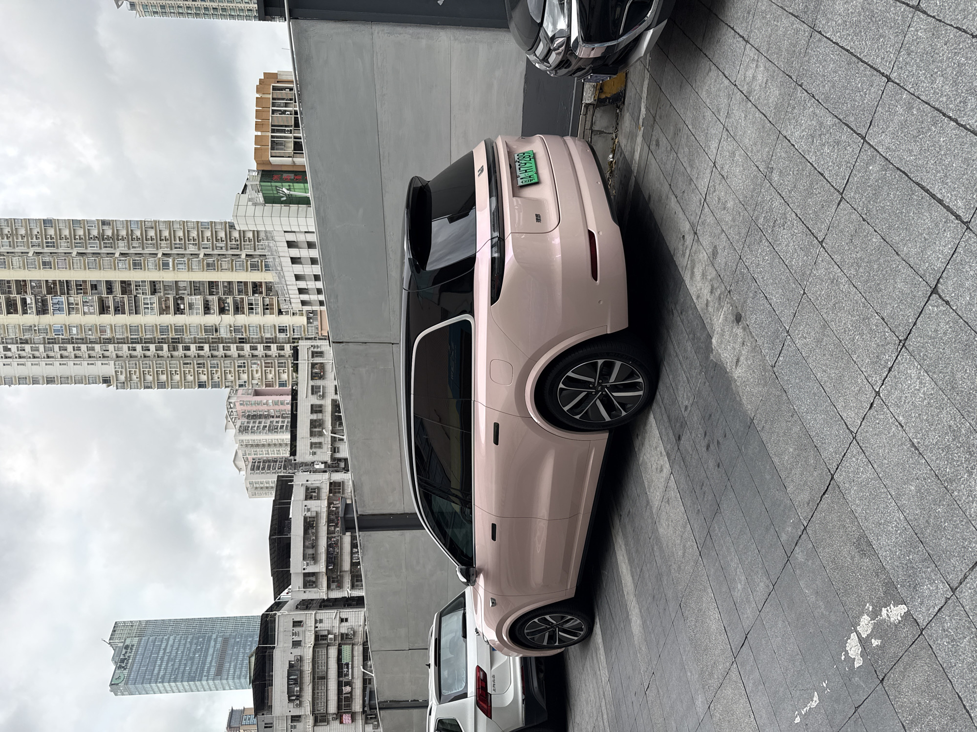 The image shows a pink XPeng G9 electric SUV parked on a gray tiled sidewalk in an urban environment. The car has a black roof and a green license plate that reads "粤B AHTP289". Behind the pink SUV, there is a white vehicle. In the background, there is a view of multiple high-rise buildings under a cloudy sky, typical of a dense urban area. The scene appears to be in the Futian District of China based on the license plate. A dark colored sedan partially obscures the rear of the pink SUV.
