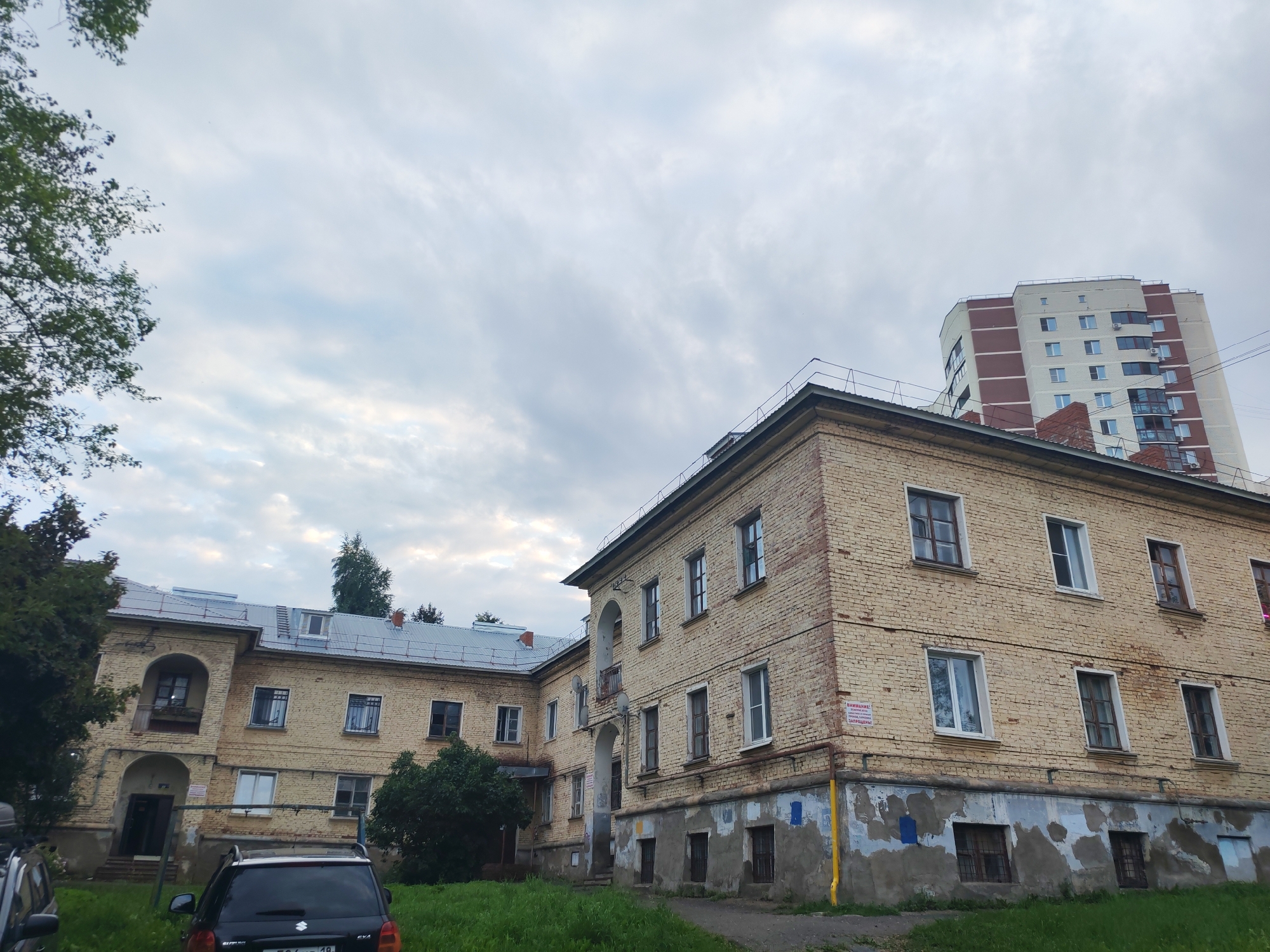 The image captures an exterior view of a multi-story brick building with an overcast sky in the background. The building appears to be older, with some signs of wear and tear, particularly at the lower levels. The brickwork is a pale yellow, and many of the windows are visible, some with barred lower panes. There are also several trees and green grass in the foreground, suggesting an outdoor setting. In the background, a taller, more modern apartment building with red and white siding is visible. A black SUV is parked in front of the brick building, and other vehicles and infrastructure, such as power lines, are also present. The overall scene suggests a residential or institutional area within a city.
The building in the foreground is located at Ulitsa Komsomolskaya, 179 in Izhevsk, Russia, which is a former dormitory of the Izhevsk Mechanical Plant.