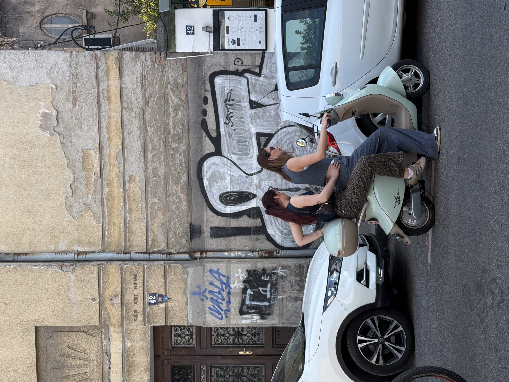 Two women are posing on a light blue scooter parked on a city street. The scooter is positioned between a white compact car and a building. The woman nearest to the car is leaning back, while the other is facing forward and holding up a cell phone, as if taking a picture. The building adjacent to the scooter has graffiti art on the side. The number "42" is visible on the building's exterior. The location is Belgrade, Serbia.