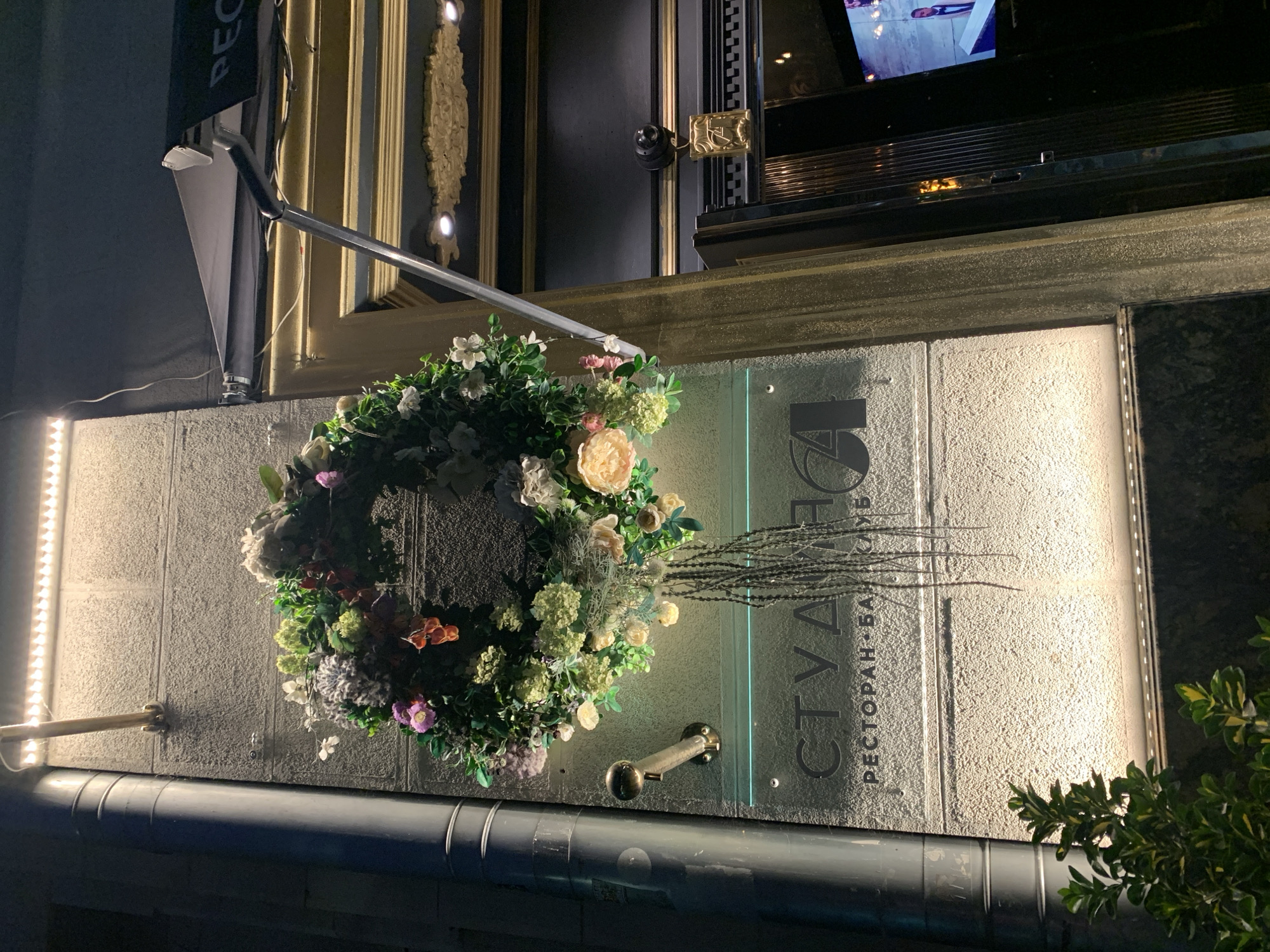 An outdoor, nighttime shot shows a floral wreath hanging on a textured stone wall in front of a building entrance. The wreath is composed of various artificial flowers and greenery in shades of white, pink, green, and purple. Below the wreath, a frosted glass sign displays the text "СТУДИЯ" in large, dark letters, with smaller text below reading "РЕСТОРАН • БАР • КЛУБ". To the left of the sign, a large, dark gray downspout runs vertically along the wall. A bright LED light strip is mounted horizontally above the wreath, illuminating the scene. To the right of the sign and wreath, the building entrance features a dark facade with decorative gold trim and a glass door. A security camera is mounted above the door, with a golden decorative plaque hanging from it. A television screen is visible inside the building. A small green plant is in the lower left corner of the frame.