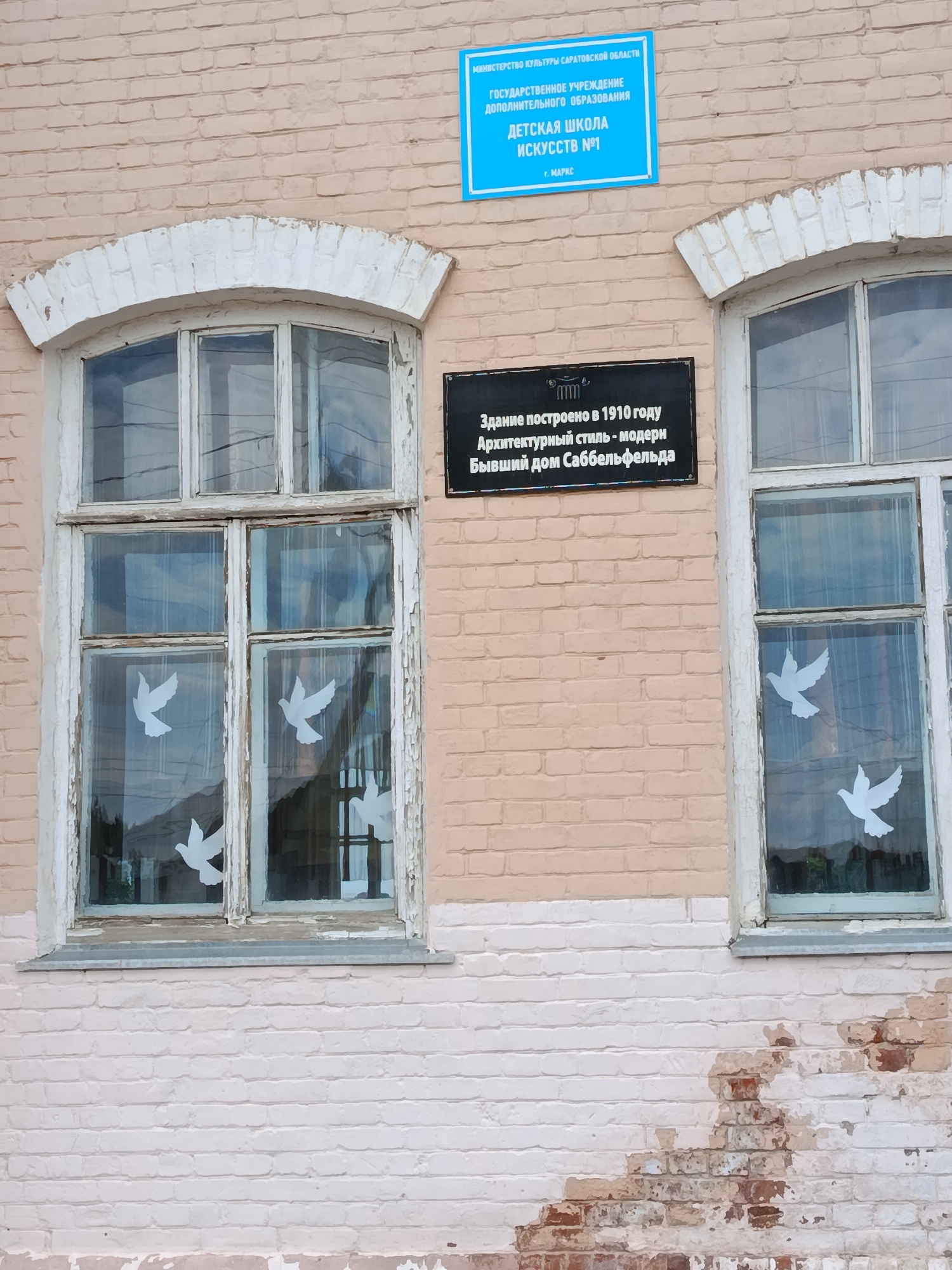 The image shows the facade of a building in the city of Marx, Russia. A blue sign is mounted on the brick wall above a window. The sign reads "MINISTRY OF CULTURE SARATOV REGION" at the top, followed by "STATE INSTITUTION OF ADDITIONAL EDUCATION" and then "CHILDREN'S ART SCHOOL NO. 1". Below this, it says "g. MARKS". To the right of this sign is a black plaque with text in Russian. This plaque states that the building was constructed in 1910 in the modern architectural style and was formerly the Sabbelfeld house.
The building has a light pink brick facade. There are two large windows visible, each with white wooden frames that appear to be aged and peeling paint. The windows have multiple panes, and paper cutouts of white doves are adhered to the glass of the left window. Reflections of the sky and trees are visible in the windows. The lower part of the brick wall shows signs of wear and damage, with some bricks exposed and discolored.