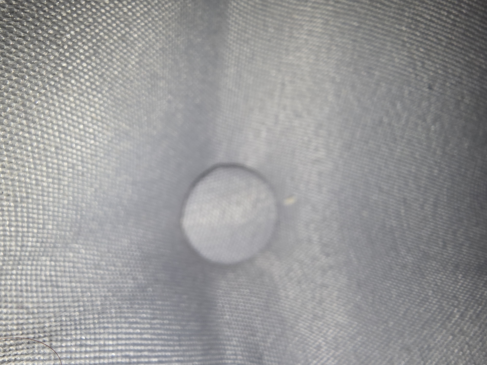 The image is a close-up, out-of-focus shot of a tufted button on a gray upholstered surface. The texture of the fabric is visible, with a grid-like pattern. There is a single dark hair visible on the left side of the image. The lighting creates highlights and shadows on the fabric. There is no discernible location in the city of Ulan-Ude, Russia, or any people or actions depicted.