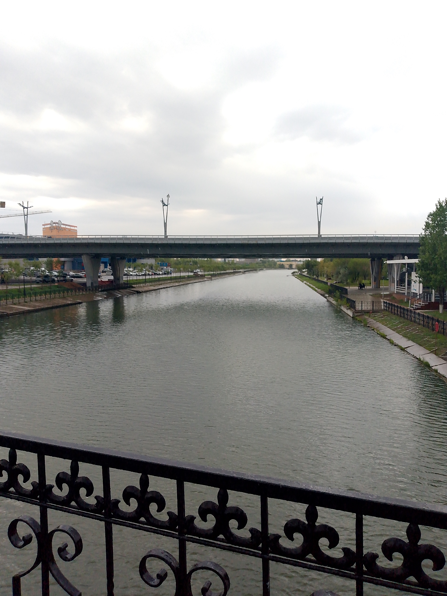 The image shows a view of the Ishim River in Astana, Kazakhstan, looking west. A large overpass bridge spans the river, supported by concrete pillars. The bridge is lined with streetlights and has a railing. On the north bank of the river, there are trees, a walkway, and a parking lot filled with cars. Further in the background, there is a building with orange siding and a crane. On the south bank, there is a white building with glass windows, a black metal fence, and more trees. The sky is overcast, and the water is calm with visible ripples. In the foreground, a black decorative metal railing is visible.
