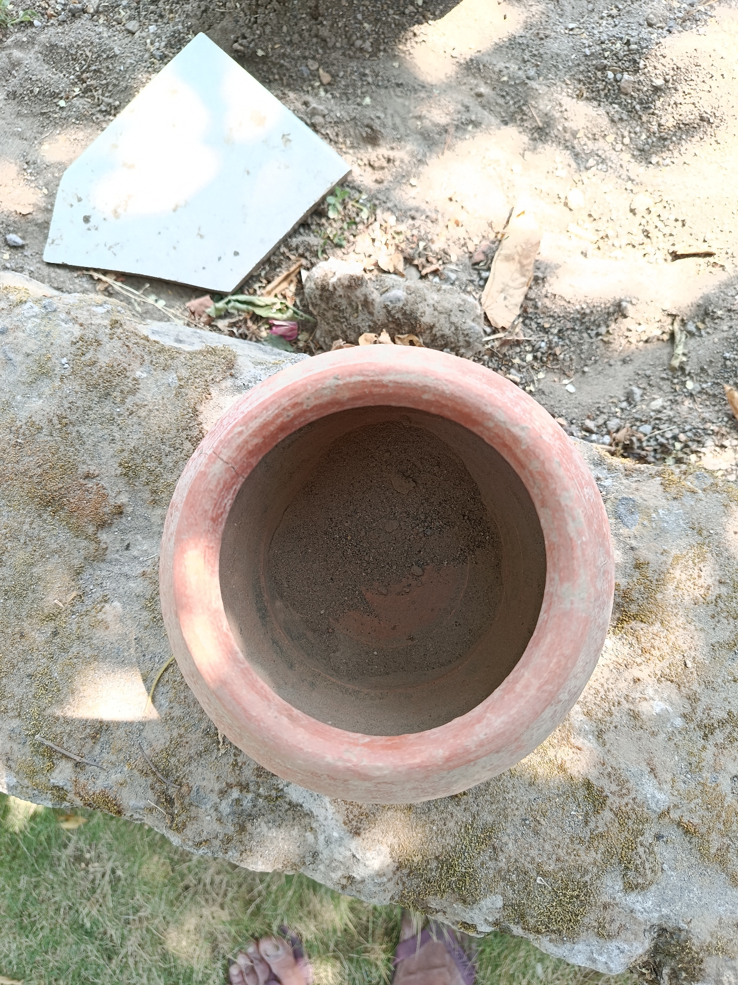 A top-down, eye-level shot captures a terracotta pot filled with dirt, resting on a moss-covered stone in what appears to be an outdoor setting in Kemiri, Indonesia. The pot is positioned centrally, its circular opening a dark void against the muted orange of the clay. Sunlight casts dappled shadows across the scene, highlighting the texture of the rough ground, scattered pebbles, and dry leaves. To the left of the pot, a white, angled tile is partially visible, lying on the dusty earth. In the foreground, the blurry impression of two bare feet indicates a person is present, though only their feet are in view, suggesting they are standing just out of frame or the camera is positioned lower than their eye level. The grass in the bottom left corner is out of focus, further contributing to the overall sense of a candid, everyday moment.