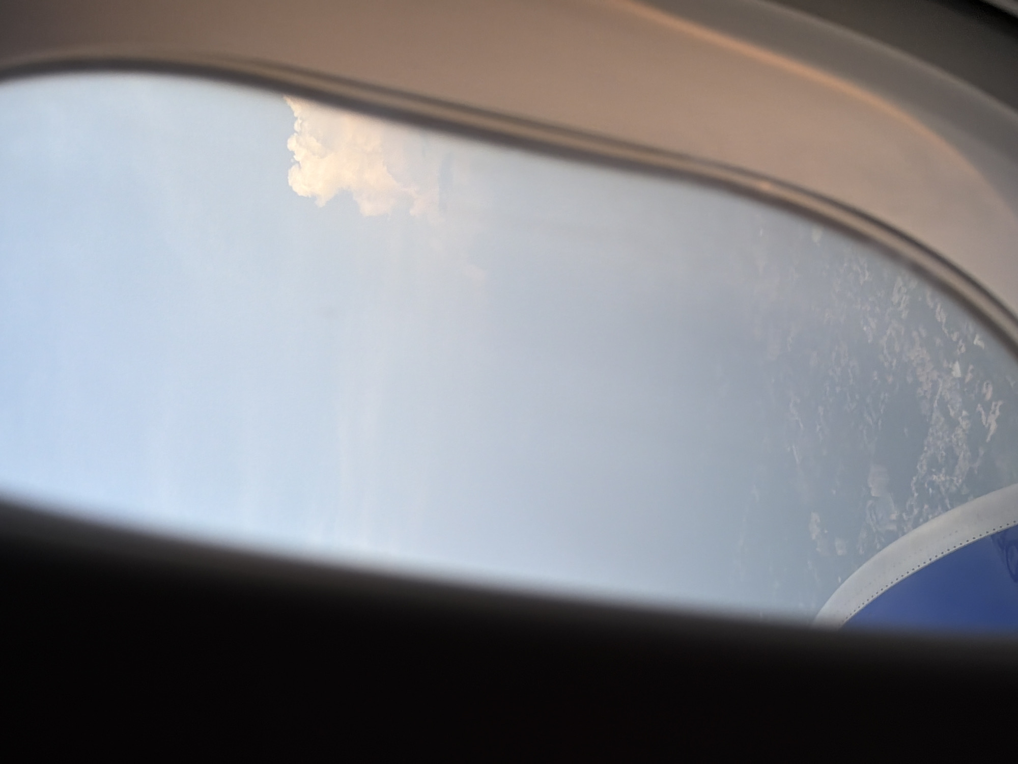 This is a photograph taken from an airplane window, looking down at the city of Nadergul, India. The view is partially obscured by the airplane's interior, including a dark, blurred section on the left and a lighter, beige frame around the window on the right.
The sky is a pale blue, with a single, fluffy white cloud visible in the upper right quadrant of the frame. Below the sky, the city of Nadergul is visible. The cityscape appears to be dense with buildings and vegetation, rendered in muted tones of green and grey due to the distance and atmospheric haze. Some structures, possibly apartment buildings or commercial complexes, can be discerned as clusters of geometric shapes. The overall impression is of a sprawling urban or semi-urban environment seen from a significant altitude.
In the lower left corner of the image, a portion of the airplane's interior is visible, featuring a curved section with a metallic rim and a blue colored surface. This part of the airplane's seat or interior structure is out of focus and appears to be part of the overall perspective from the window seat.
There are no people or discernible actions taking place in the image. The scene is a static, aerial view of a landscape.