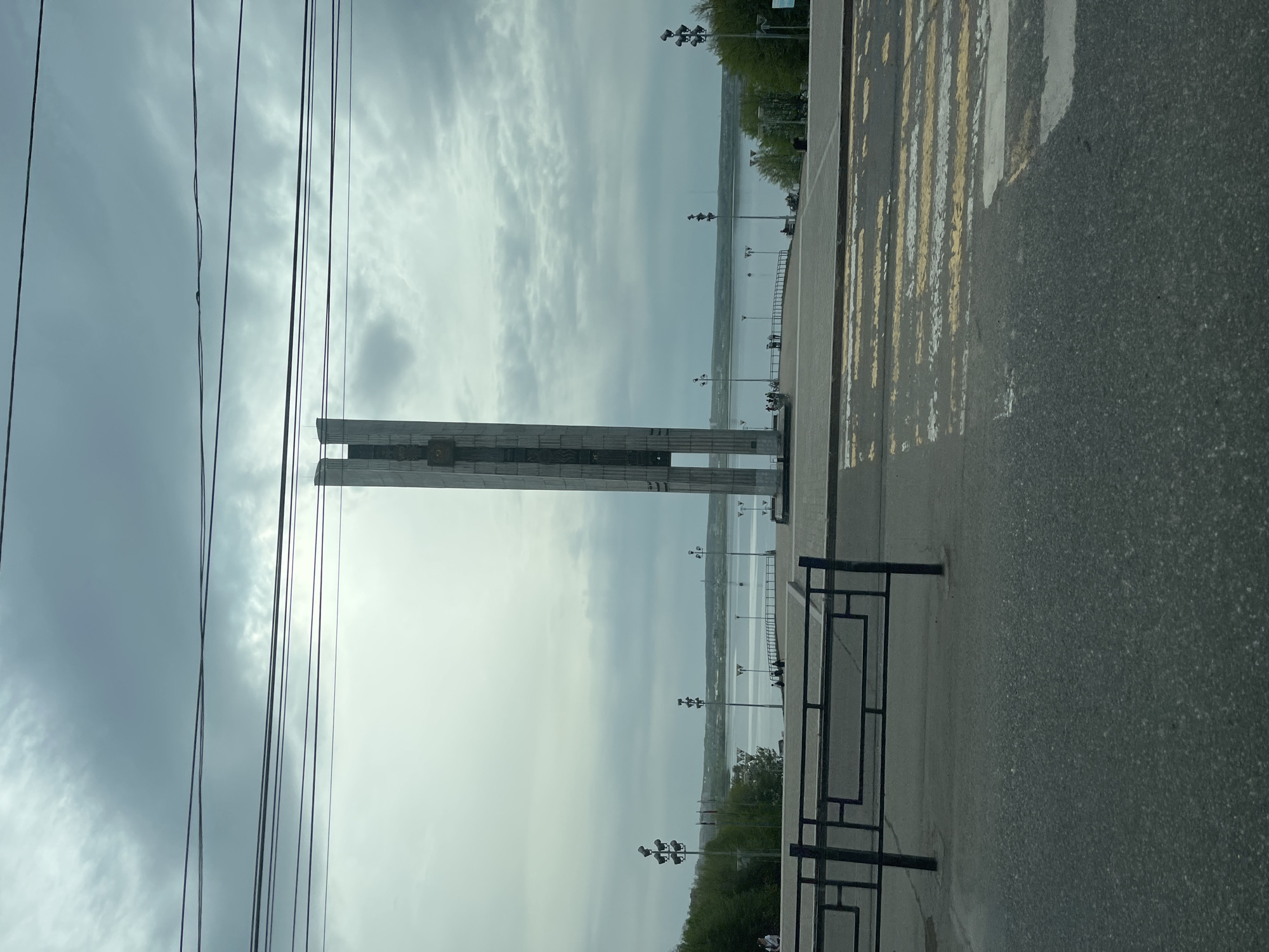 The image shows a vertical view of a cityscape in Izhevsk, Russia. A tall, rectangular monument stands prominently in the foreground, with power lines visible to its left. In the background, a body of water stretches out to the horizon under a cloudy sky. The waterfront is lined with street lamps and greenery. To the right of the monument is a crosswalk and a section of roadway. A black metal fence is also visible along the pavement.