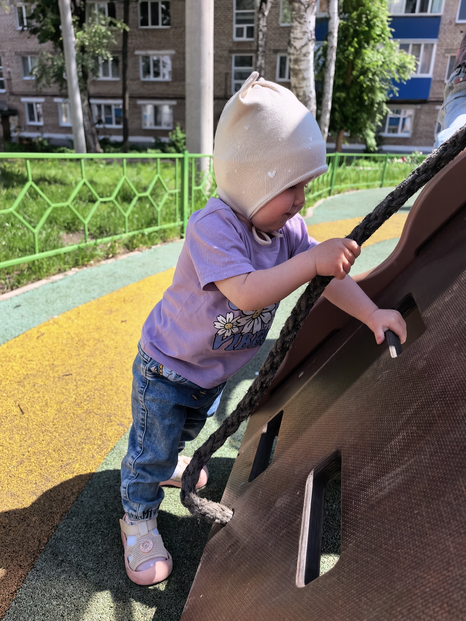 In Sterlitamak, Russia, a young child, possibly a toddler, is on a playground. The child is wearing a light-colored bonnet adorned with small hearts, a lavender t-shirt with a daisy print, blue jeans, and pink sandals. The child is gripping a thick rope with their hands and reaching for a handhold on a brown climbing structure. The playground features a yellow and green surface. In the background, there are apartment buildings and a green fence. The lighting suggests it is daytime, with shadows cast on the ground.