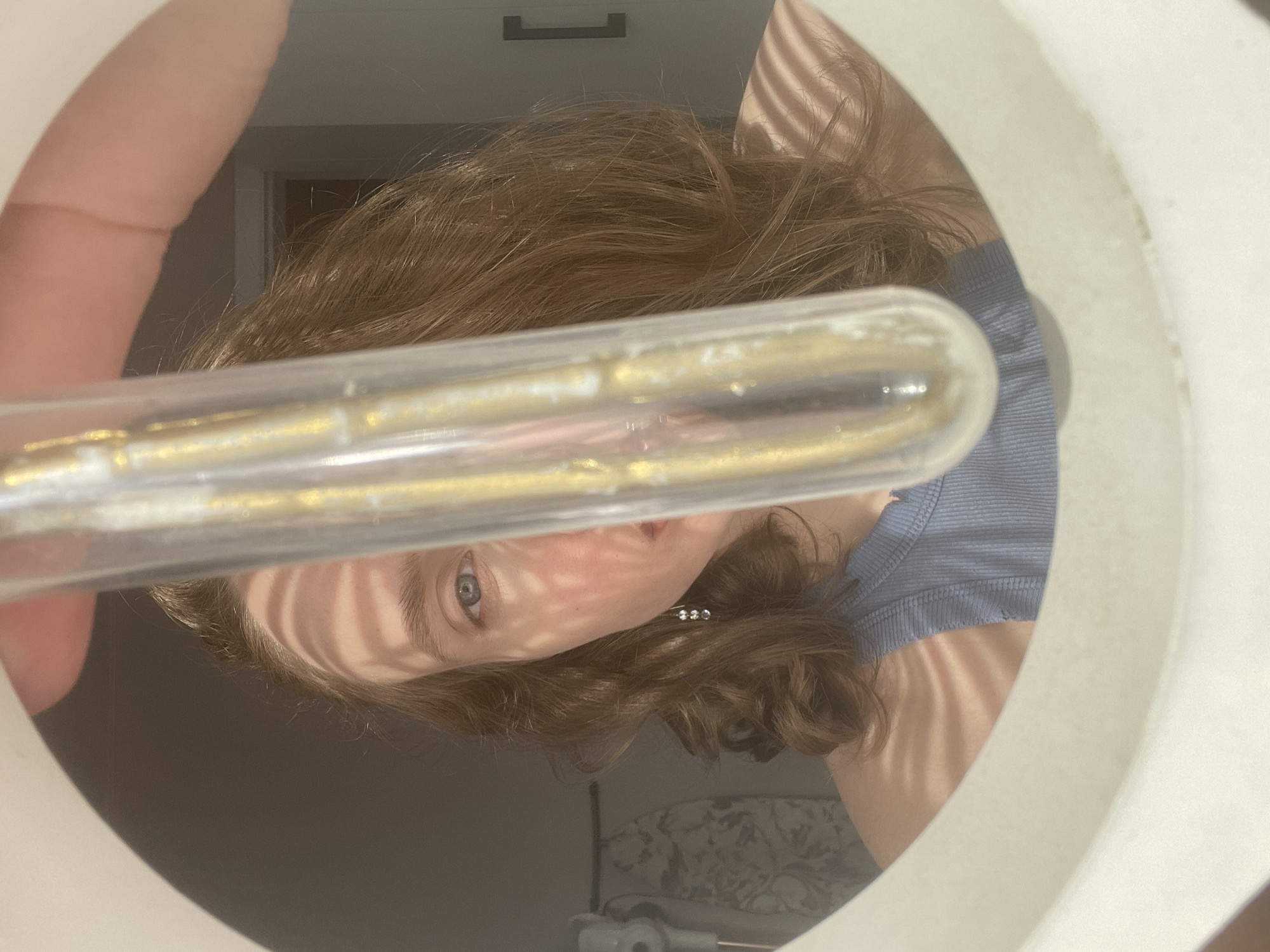 A person with light brown hair, blue eyes, and a pale complexion is holding a clear tube filled with a golden substance in front of a white circular object, possibly a light fixture or a mirror frame. The person is wearing a light blue shirt. The background is blurry, but a door and a toilet can be partially seen. The location is impossible to verify based on the image content alone.