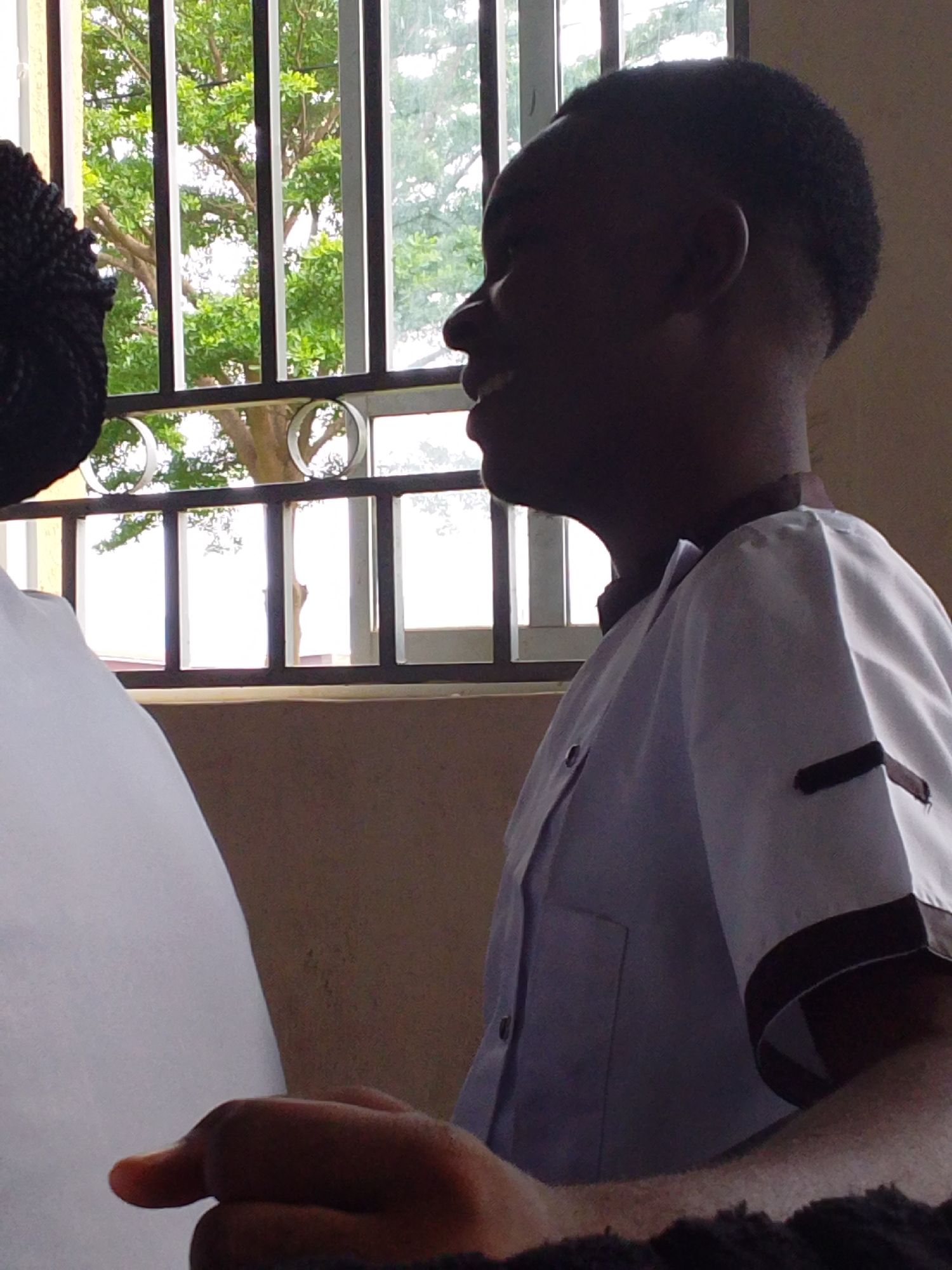 The image is an indoor, medium shot of two young Black individuals positioned behind a barred window. On the left, a person's head and shoulder are visible, with dark hair styled in braids. To the right, a boy or young man wearing a light-colored school uniform with dark trim is shown in profile, looking out the window and smiling. The window features a black metal grate and offers a view of green foliage and trees, suggesting an outdoor environment beyond the building. The interior wall behind the subjects is a neutral beige color. The lighting suggests a daytime setting, possibly late morning or early afternoon, with natural light filtering in through the window. The specific location in Kuson, Nigeria, is not determinable from the image.
