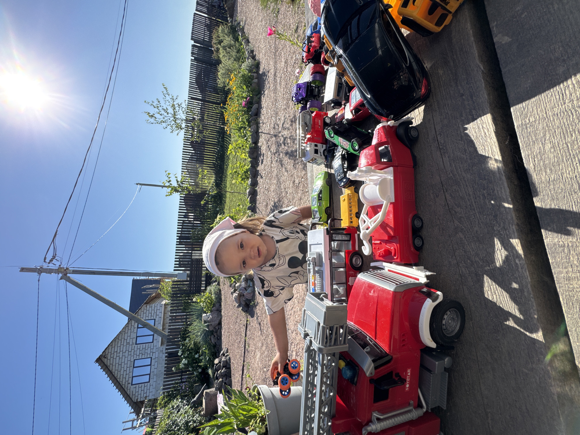 A young girl with a pink headscarf is playing with a collection of toy vehicles on a wooden surface outdoors, possibly in Kempelevo, Russia. The girl is wearing a patterned t-shirt and is reaching for a toy car. The toys include various types of cars, trucks, and a fire engine. Behind the girl, a house under construction and a wooden fence are visible, along with some plants and flowers. The sun is shining brightly, casting shadows on the wooden surface.