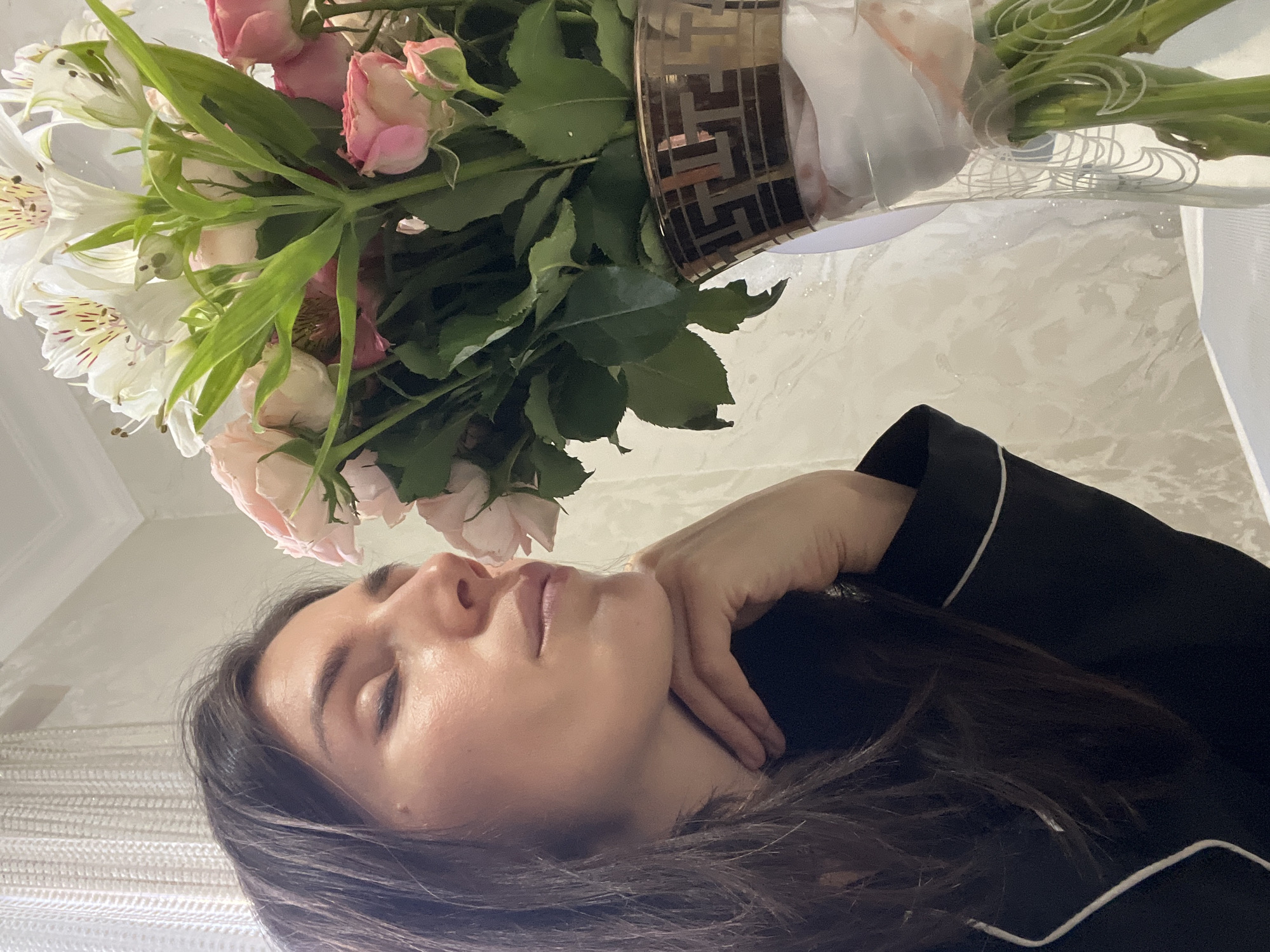 A low-angle, rotated close-up captures a serene indoor scene featuring a woman and a bouquet of flowers. The woman, positioned in the lower right, has dark brown hair and is seen in profile with her eyes closed or looking down, conveying a calm or contemplative mood. Her right hand gently touches her neck. She wears a dark top, possibly a pajama shirt, with distinctive white piping along the edges.
Above and to the left of the woman is a bouquet composed of light pink roses or rosebuds and white lilies (Alstroemeria) characterized by their spotted inner petals. The stems are gathered by a transparent material adorned with white, intricate swirling patterns. The flowers are held in a container featuring a gold-colored band with a repeating geometric pattern.
The background is a light-colored, subtly textured surface, likely a wall or ceiling. A sheer white fabric, possibly a curtain, is visible on the left. The soft, diffused lighting contributes to the peaceful atmosphere. The image depicts a quiet moment of relaxation or appreciation of the flowers in an intimate, domestic setting. No visible text is present. The location context is Kaspiysk, Russia.