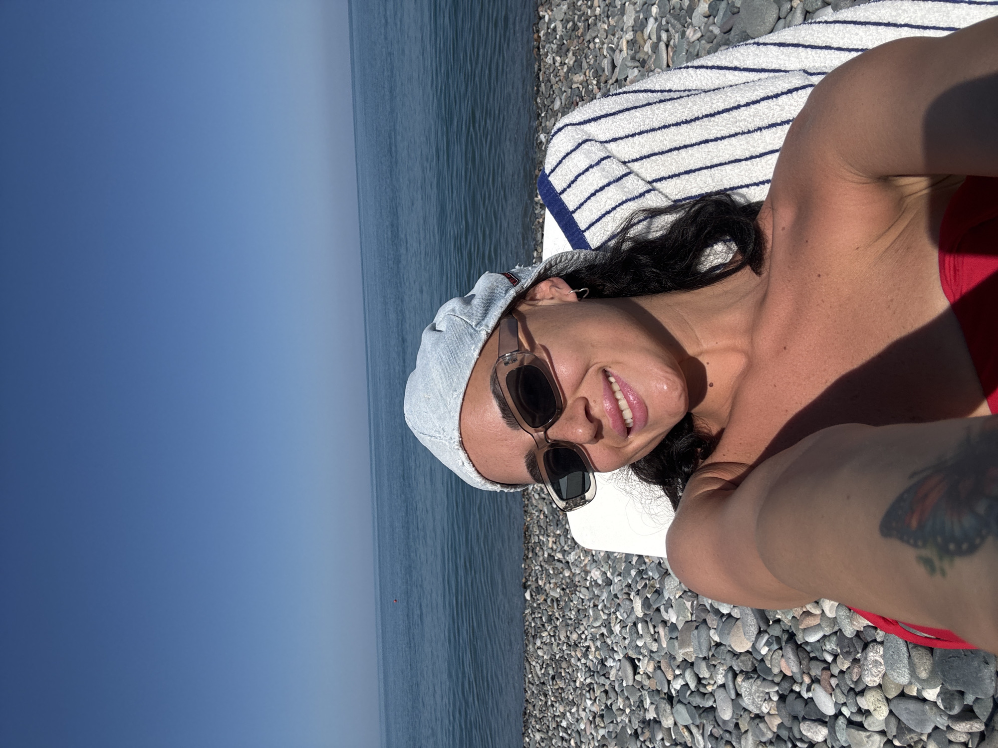 A woman is lying on a white lounge chair on a pebble beach with the sea in the background. She is wearing a light blue denim cap, sunglasses, and a red bikini top. The sea is calm and blue under a clear sky. The beach is covered in smooth, grey pebbles. A striped towel with blue and white stripes is draped over the back of the chair. The woman is smiling and looking at the camera. The overall scene suggests a relaxed day at the beach in Batumi, Georgia.