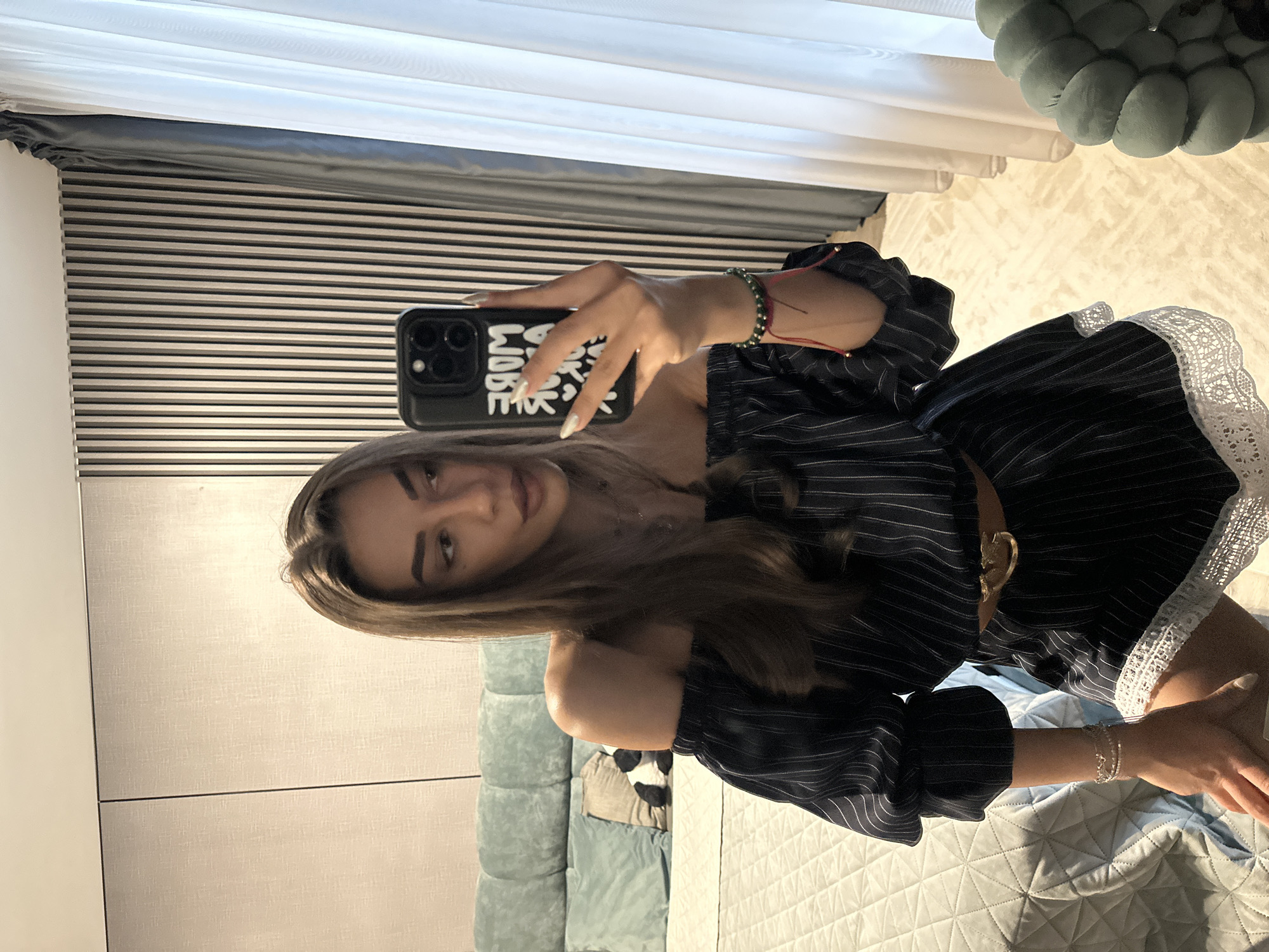 A young woman with long, brown hair and light skin is taking a selfie in a bedroom. She is wearing an off-the-shoulder black and white pinstripe dress with white lace trim. A brown belt with a gold buckle cinches her waist. She has on bracelets on her left wrist, one with green beads and a red string, and another with multiple delicate silver bands. Her nails are painted white. In her right hand, she holds a black smartphone with a case that has white lettering. The background shows a bed with a textured, light-colored comforter and a teal upholstered headboard. Behind her, there are vertical wooden slats on the wall and sheer white curtains. The scene appears to be indoors, in a residential setting, possibly a bedroom in a hotel or apartment. No specific location within the city of Sofia, Bulgaria, can be identified from the visual information provided.