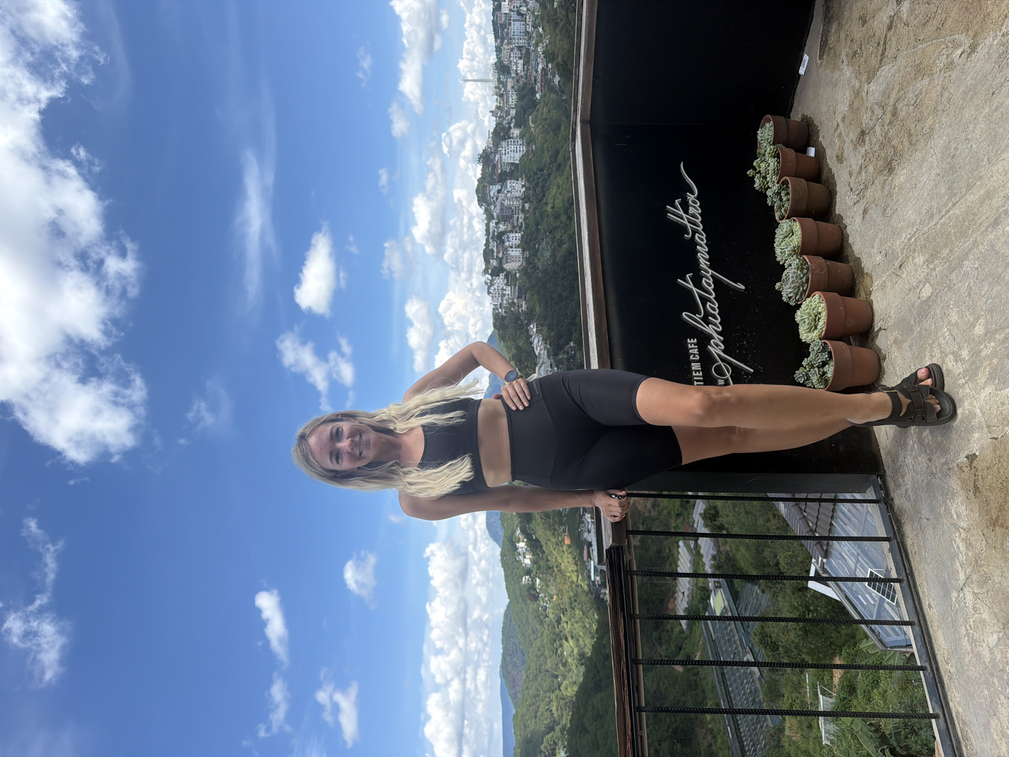 A blonde woman stands on a balcony overlooking a cityscape in Da Lat, Vietnam. She is wearing a black sports bra and black biker shorts. Her left hand is on her hip, and her right hand rests on a railing. In the background, a town is nestled amongst rolling green hills under a partly cloudy blue sky. The balcony has a concrete floor and a black metal railing. Along the back wall of the balcony, there are several potted succulents and a neon sign that reads "TIEM CAFE Sophiaymattra".