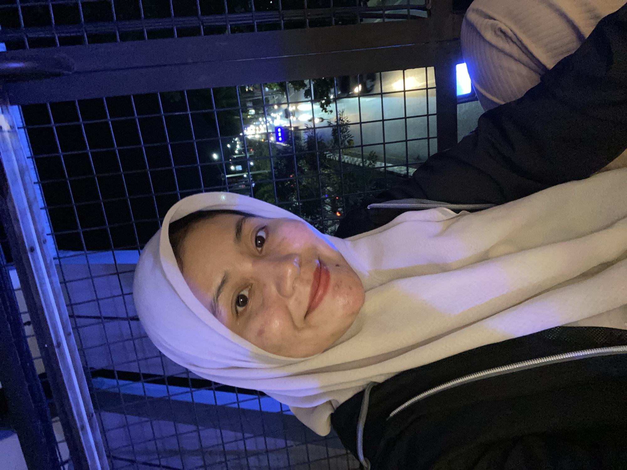 The image shows a close-up selfie of a woman wearing a light-colored hijab and a black jacket. She is looking at the camera and smiling. In the background, through a metal grate, a street scene at night is visible with car headlights and streetlights illuminating the road and surrounding trees. The location is identified as Banjarbaru, Indonesia. The overall ambiance suggests an outdoor setting at night, possibly an elevated viewpoint overlooking a city street.