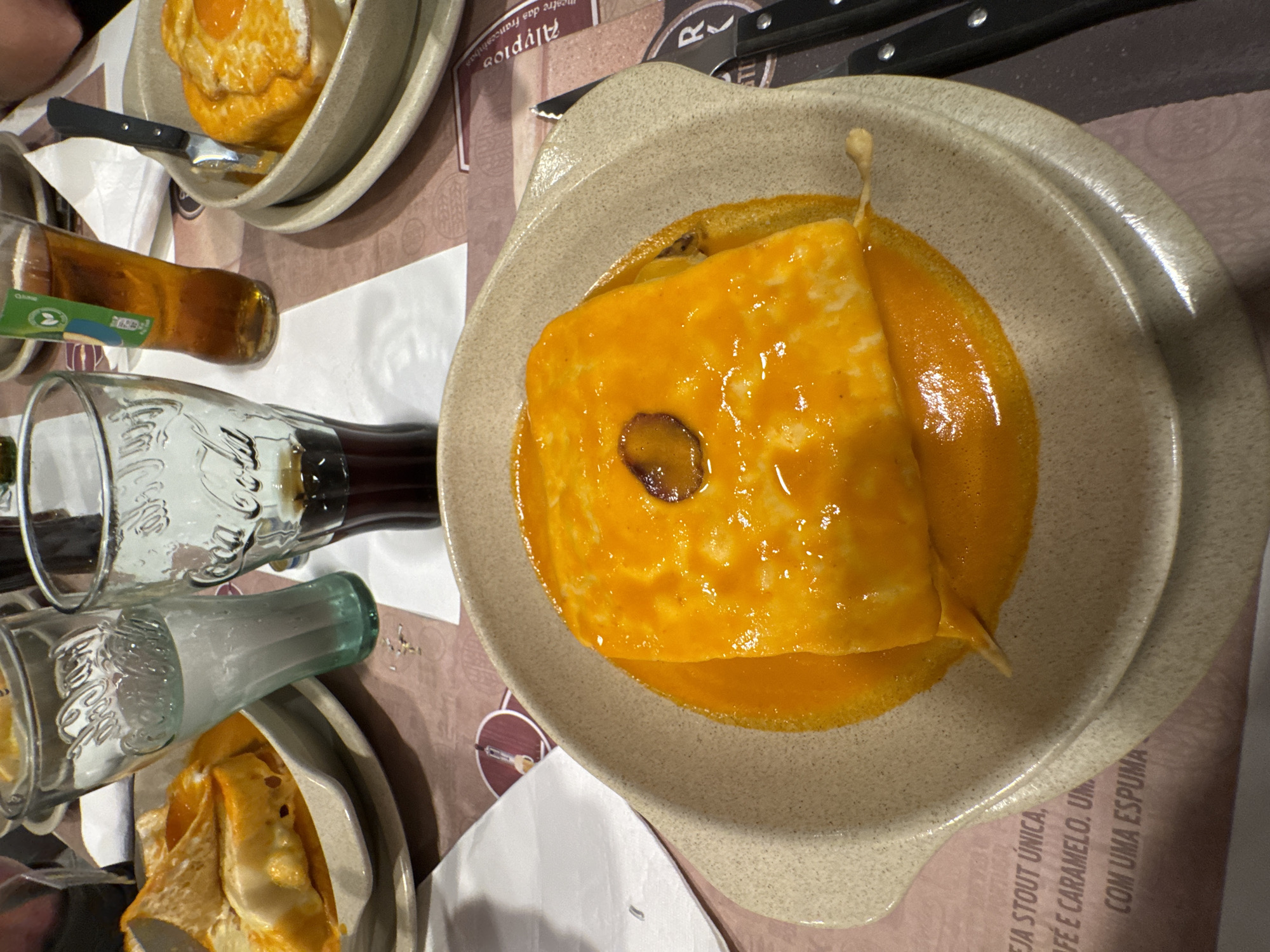 A high-angle shot captures two plates of Francesinha sandwiches on a table, accompanied by a Coca-Cola bottle and what appears to be an alcoholic beverage. The plates are light brown and speckled, and they are set against a background of a printed table covering that contains text, including the word “Alyprio.” The plate on the right contains a Francesinha sandwich almost completely covered in a bright yellow sauce. There's a dark circular dot on the surface of the sandwich, perhaps a topping or a part of the preparation. The left plate is partially visible and also contains a Francesinha sandwich. The Coca-Cola bottle is transparent glass, showing the liquid inside. The alcoholic beverage bottle is amber in color. The picture was taken in the city of Bragança, Portugal.