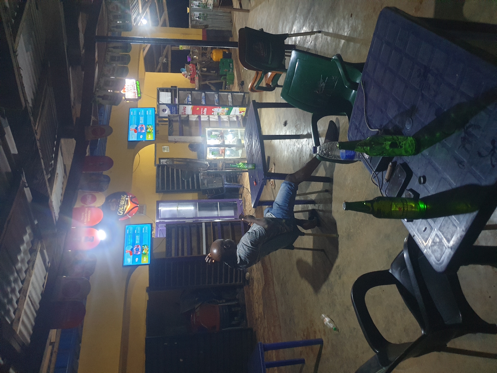 The image depicts an outdoor seating area, possibly a bar or restaurant, in Anilelerin, Nigeria. The setting appears rustic, with a corrugated metal roof supported by visible beams. Yellow walls are visible in the background, along with several shelves stocked with various items, including what appear to be beverages. Two flat-screen televisions are mounted on the wall, displaying colorful content.
In the foreground, a man is seated on a chair, leaning back and looking at his phone. He is wearing a striped t-shirt and blue jeans. In front of him, a dark-colored table holds two beer bottles, one upright and another on its side, along with a phone and a cigar. Another table with a blue top is partially visible on the left, with a discarded plastic water bottle on the ground nearby. Several other chairs, including two plastic ones in green and orange, are scattered around the area. The lighting is dim, suggesting it is evening or night. The overall atmosphere suggests a casual, possibly relaxed, social gathering space.