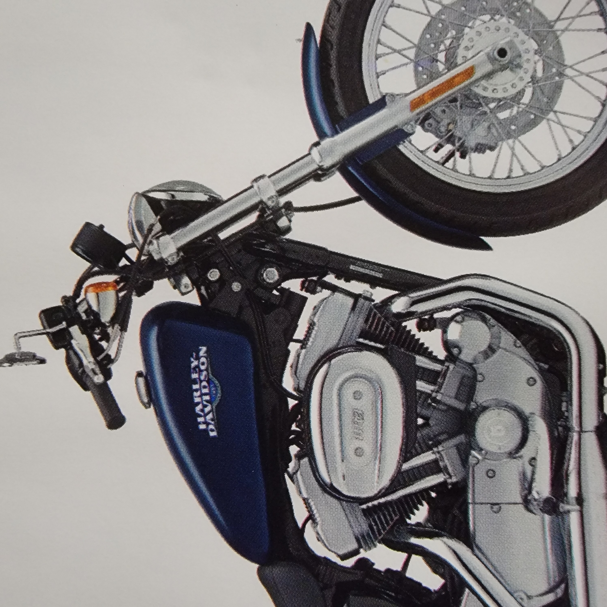 A close-up, eye-level shot shows the front portion of a dark blue Harley-Davidson motorcycle. The motorcycle is shown against a plain white background, with no environmental context provided. The focus is on the fuel tank, handlebars, front suspension, and wheel. The fuel tank features the Harley-Davidson logo in silver. The handlebars are black with chrome accents. The front wheel is a spoked wheel with a disc brake and caliper. The engine is partially visible, with chrome and black components. An orange reflector is visible on the front suspension.