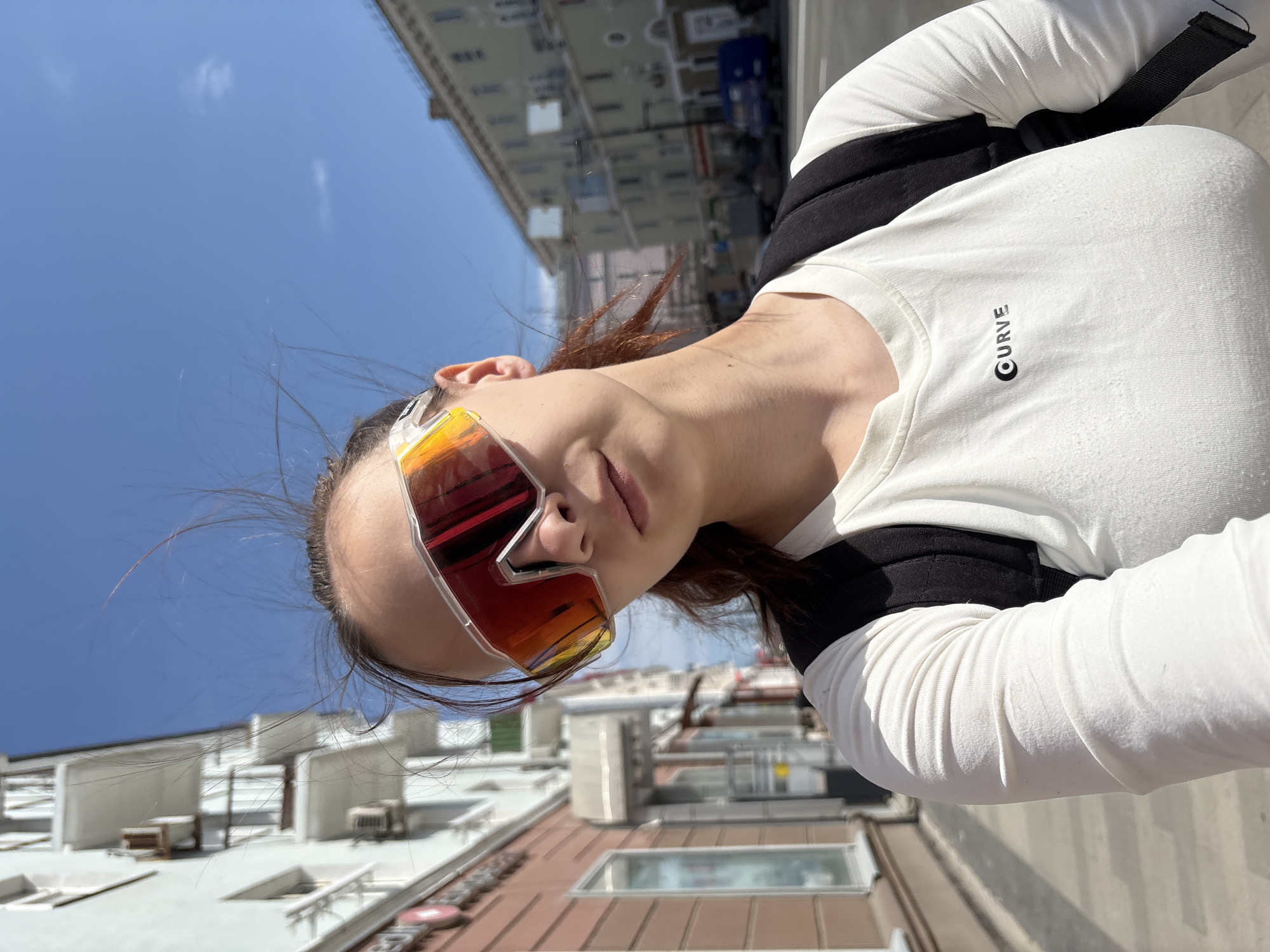 A woman with sunglasses stands on a rooftop in Perm, Russia, with a clear blue sky and surrounding buildings in the background. She is wearing a long-sleeved white shirt with a black strap visible over her shoulder. The city is reflected in the lenses of her sunglasses.