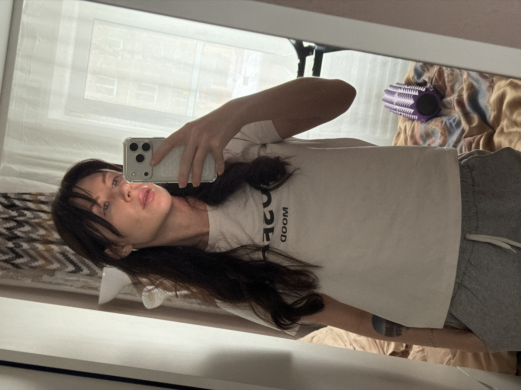 A female subject is captured in a mirror selfie within an indoor residential setting in Perm, Russia. The subject, with long dark hair and bangs, is wearing a light-colored short-sleeved t-shirt displaying the inverted word "DOOM" and grey sweatpants with a white drawstring. She holds a light-colored smartphone with a clear case and three prominent rear cameras, directing it towards her reflection. A partial, dark-colored tattoo is visible on her left upper arm. The background, reflected in the mirror, reveals a window looking out onto an urban environment with buildings and what appears to be an overcast sky. Inside the room, a bed with a multi-colored duvet (featuring hues of brown, blue, and pink) is visible, upon which a purple, textured foam roller rests. Additionally, a white standing lamp with a bowl-shaped shade and patterned curtains with a zig-zag design are discernible.
