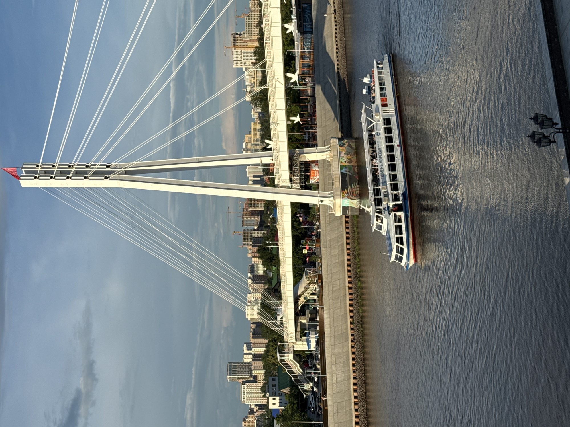 This is an aerial, rotated photograph of the Bridge of Lovers in Tyumen, Russia. A white cable-stayed bridge dominates the left side of the frame, with numerous white cables fanning out from a central tower against a bright blue sky with wispy clouds. A small red flag is visible at the top of the bridge's structure.
Below the bridge, a city skyline with numerous buildings and construction cranes is visible on the left bank. A wide, dark body of water, likely the Tura River, occupies the right side of the image. A long, white passenger boat with multiple decks and windows is traveling on the river, close to the embankment. The boat has red trim and appears to be carrying passengers. The embankment is made of concrete and brick. Two ornate streetlights stand on the right side of the embankment. The lighting suggests it is late afternoon or early evening, with long shadows cast by the bridge and streetlights. The water is rippled, indicating a slight breeze. The overall mood of the image is peaceful and scenic, showcasing urban infrastructure and leisure activity in a city setting. The boat has text on its side that reads "ТЮМЕНЬ МОРСКАЯ" (Tyumen Morskaya).