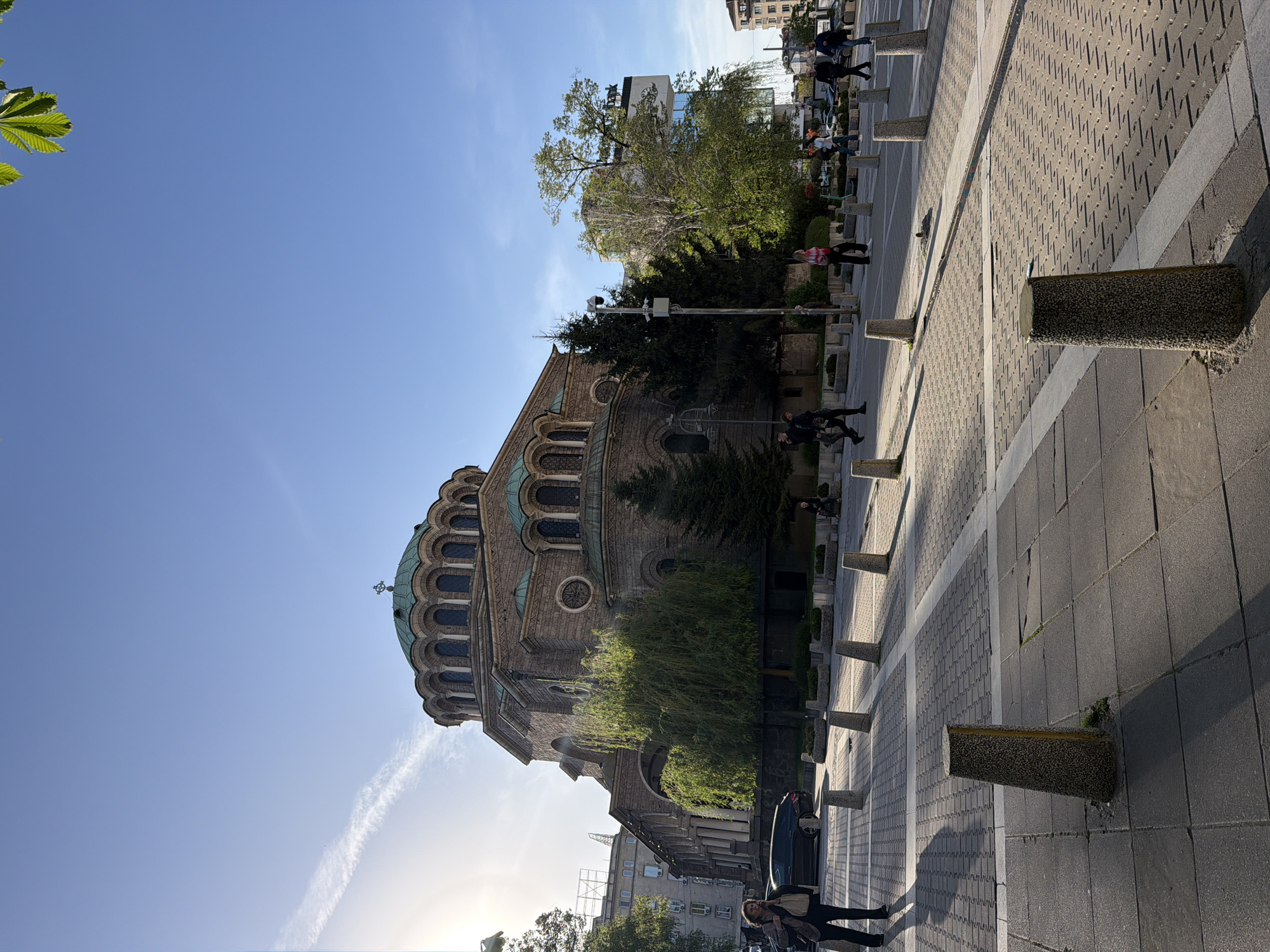 This image captures a vibrant urban street scene in Sofia, Bulgaria, characterized by a prominent church, a pedestrianized area, and various people.
The most striking subject is a large, stately church on the left, likely an Eastern Orthodox church given its architecture. It features a robust stone and brick facade, numerous arched windows, and distinctive green-patinated domes, with a large central dome surrounded by smaller ones, all topped with crosses. A substantial, lush green tree with weeping branches stands in front of the church, partially obscuring its lower section.
On the right side of the image, a wide paved area serves as a walkway or public square. It is laid with different shades of grey paving stones, some featuring a textured diagonal pattern. A series of short, dark, cylindrical bollards line the edge of this paved space.
Several people are visible throughout the scene. In the foreground-right, a woman in a light jacket and dark clothing stands near a parked dark car, looking towards the church. Further down the paved area, other individuals are walking or standing, including one person wearing a distinctive plaid shirt. More people are discernible in the distance on the far left.
The time of day appears to be late afternoon or early morning, as indicated by the low sun, which is casting long shadows across the paved ground from the bollards and people, and creating a lens flare near the bottom left of the frame. The sky is a clear, bright blue with a few faint, wispy contrails. Green foliage, including large leaves in the upper-left corner, adds natural elements to the urban setting. In the background on the right, behind some trees, a modern building is visible with blurry, indistinct Cyrillic text that is difficult to decipher. The overall impression is a sunny, ordinary day in a bustling city.