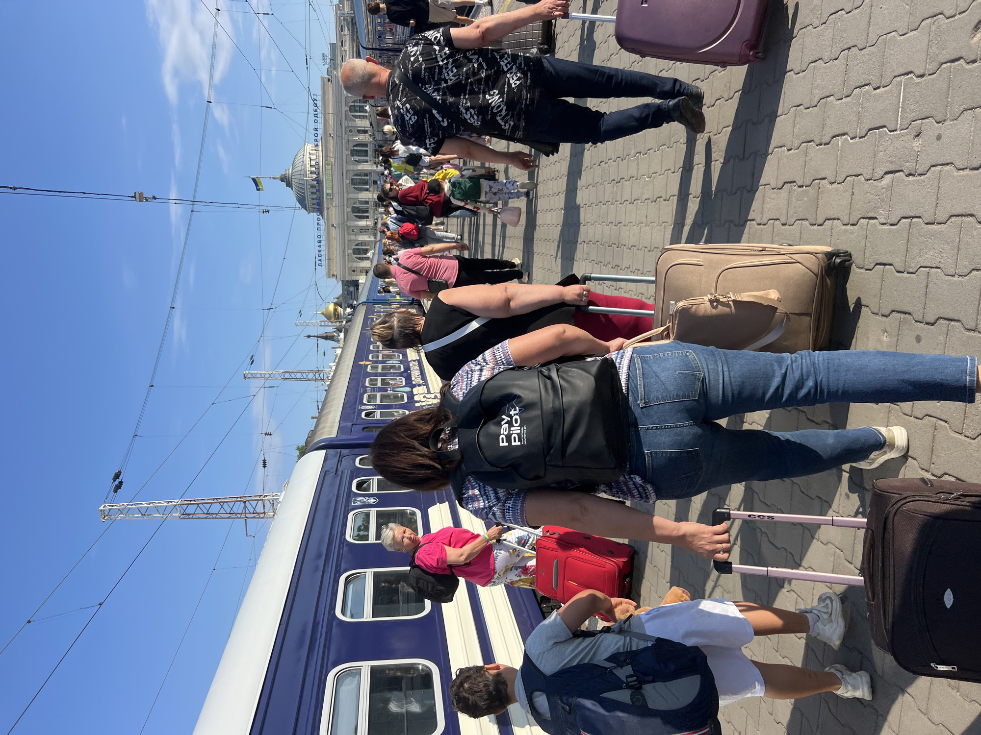 This is a full-shot, eye-level, outdoor vertical photograph taken on a sunny day at the Odesa Railway Station in Odesa, Ukraine. In the foreground, several people are walking with luggage on a paved train platform. A blue and white train is visible on the left side of the frame. In the background, the Odesa Railway Station building, with its distinctive dome, is prominent under a clear blue sky. The station features a large sign that reads "ЛАГАСКАВО ПРОСУЄМО ГЕРОЇ ОДЕСИ!", translating to "WE WELCOME THE HEROES OF ODESA!". A Ukrainian flag flies atop the dome. The scene depicts a typical moment at a busy train station, with travelers preparing to depart or arrive.