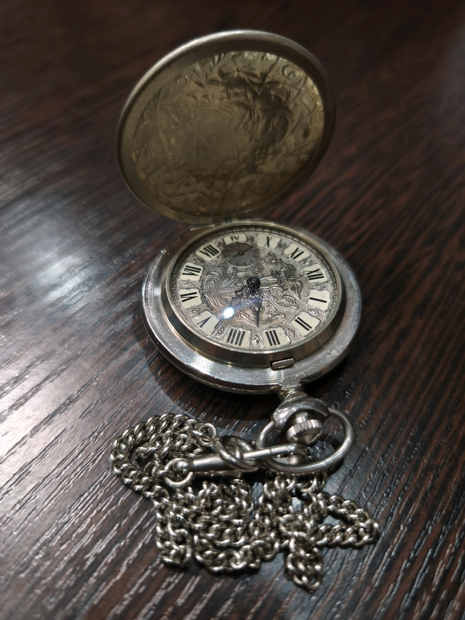 An open, ornate, silver-toned pocket watch with a metal chain rests on a dark wooden surface. The watch face displays Roman numerals and intricate detailing. The lid of the watch, also open, features decorative etching. The background is a dark, textured wooden surface. There is no indication of the city of Karachayevsk, Russia, or any people or actions within the image.
